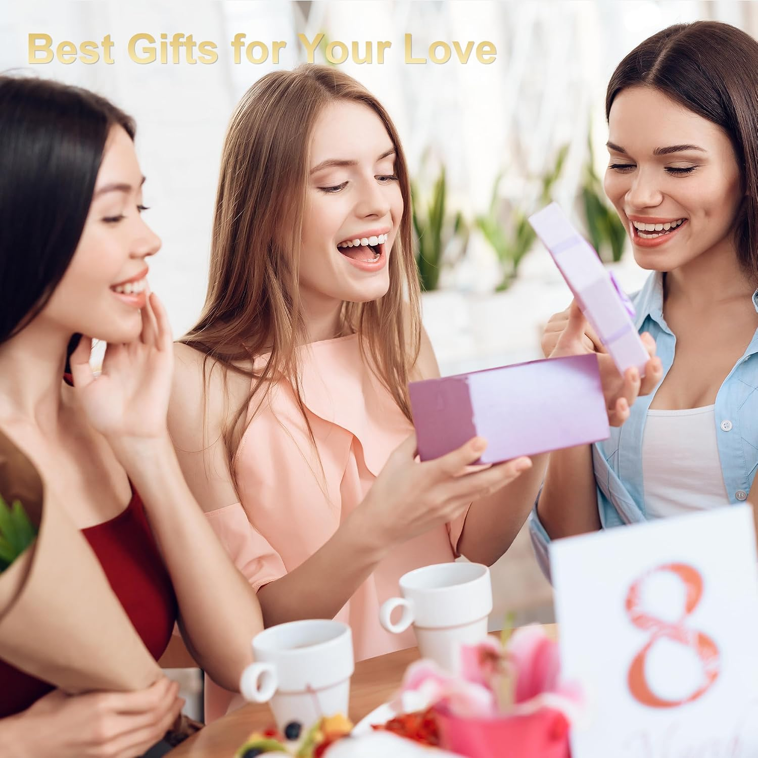 Drei lächelnde Frauen öffnen an einem Tisch eine lila Geschenkbox mit Kaffeetassen und Blumen.