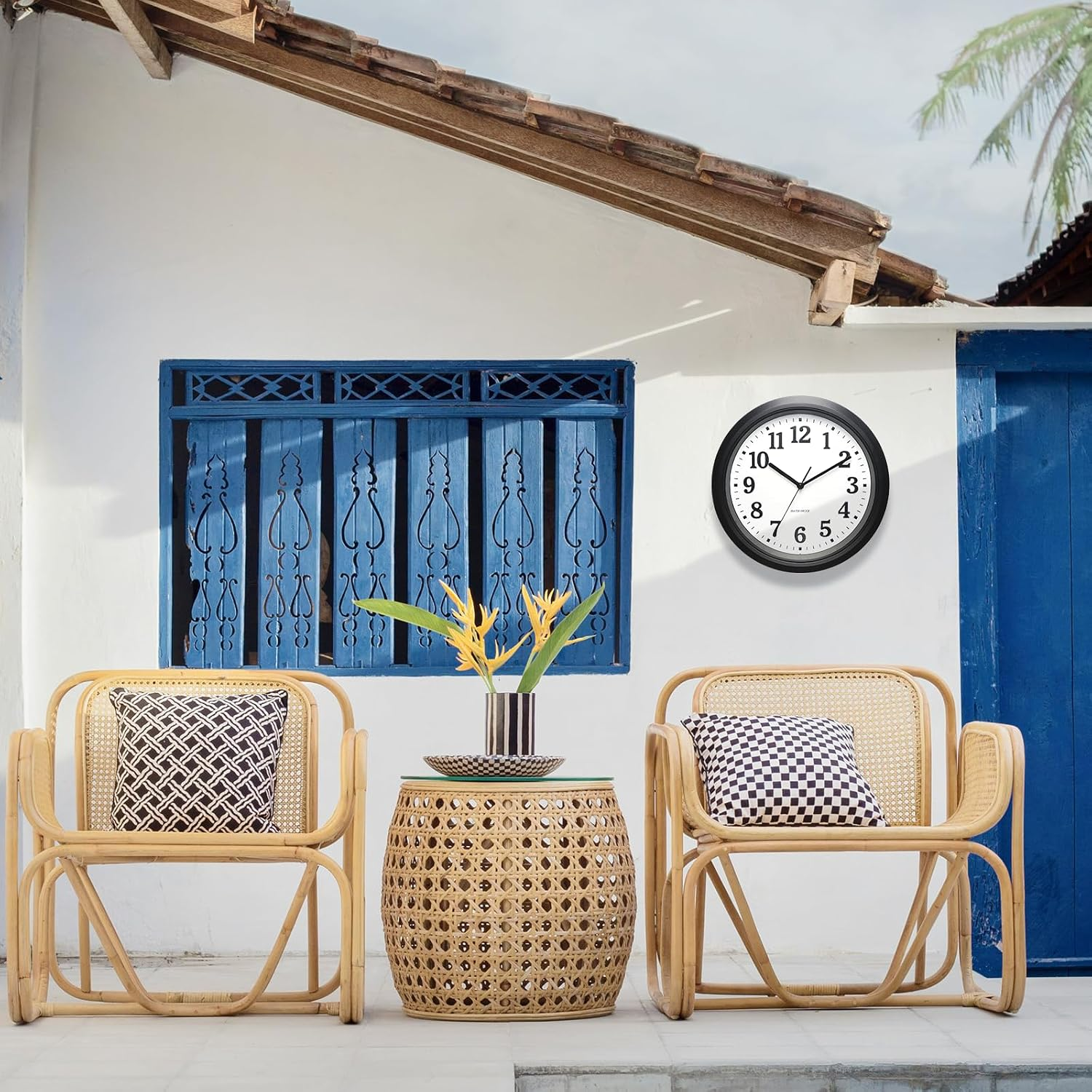 Zwei Bambusstühle, ein kleiner Tisch mit gelben Blumen, eine Uhr an der weißen Wand und ein blaues Fenster.