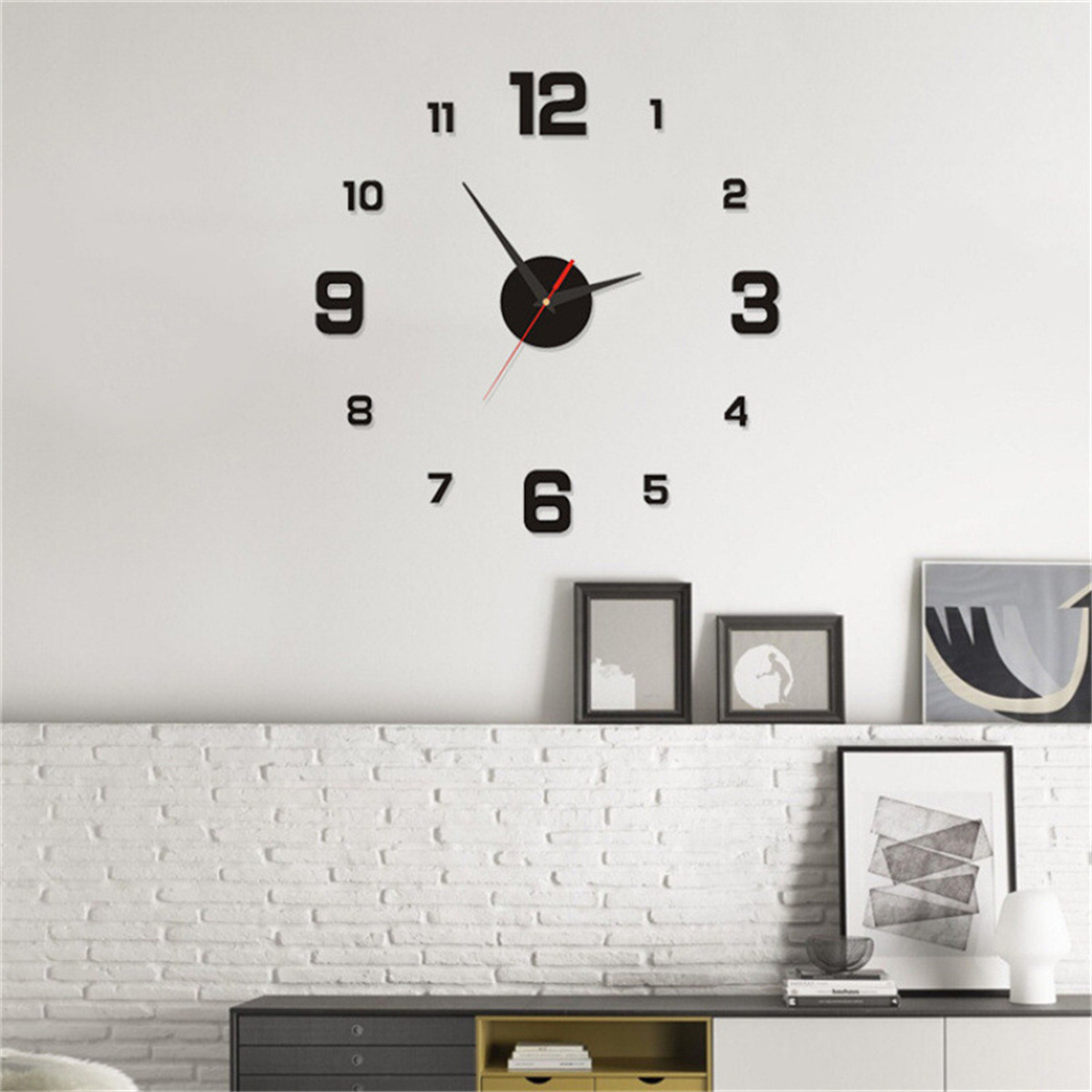 Eine große schwarze Wanduhr ohne Rahmen hängt über einer weißen Backsteinwand, darunter stehen gerahmte Bilder.