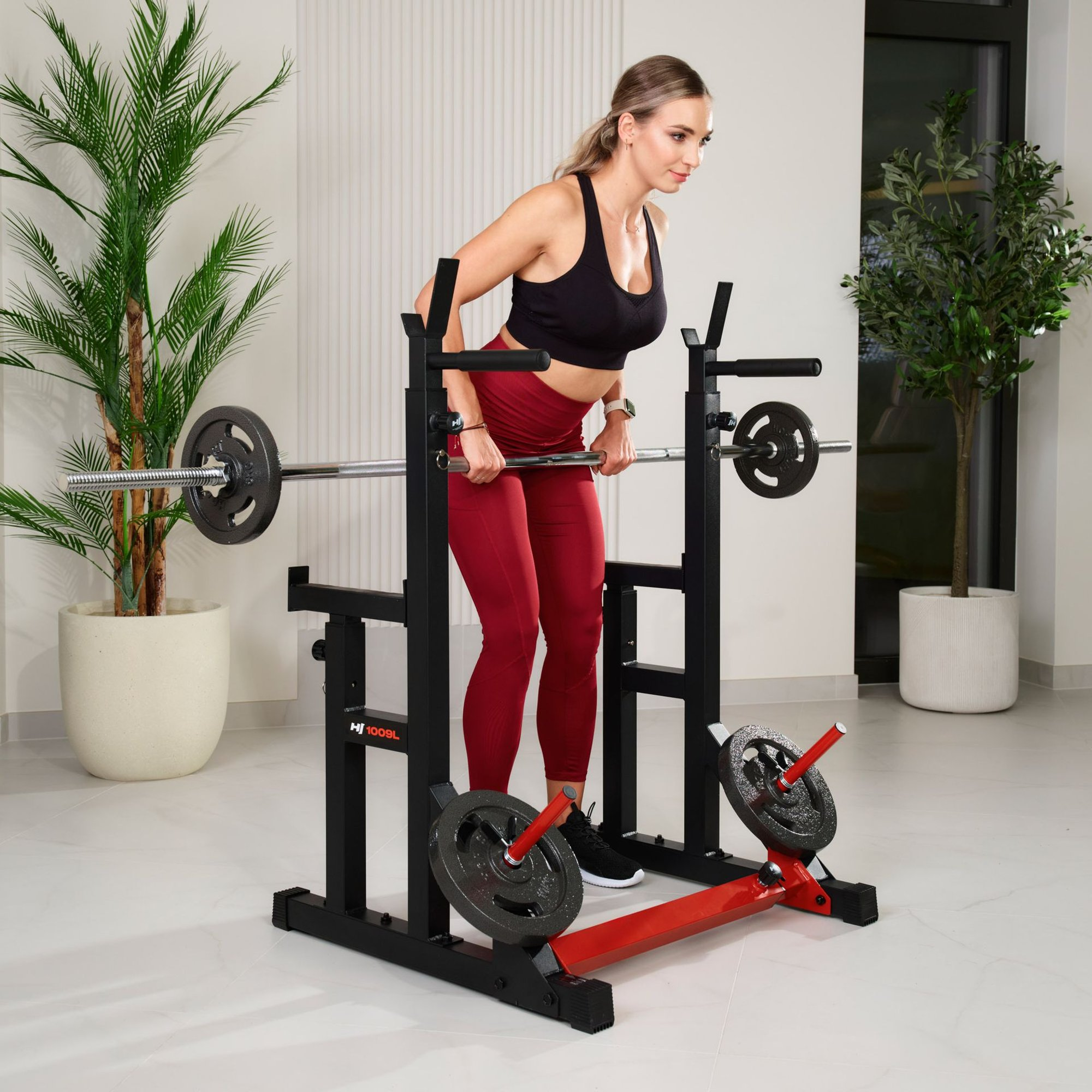 Eine Frau im schwarzen Sport-BH und roten Leggings trainiert mit einer Langhantel im Rack, umgeben von Topfpflanzen.