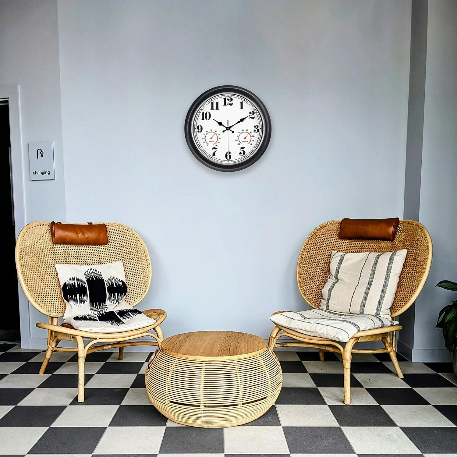 Zwei Rattansessel mit Kissen, ein runder Tisch und eine Uhr an der grauen Wand vor schwarzweißem Fliesenboden.