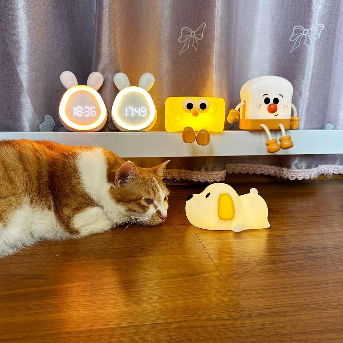 Eine orange-weiße Katze schaut auf eine kleine Hundelampe, während im Hintergrund verschiedene Nachtlichter stehen.