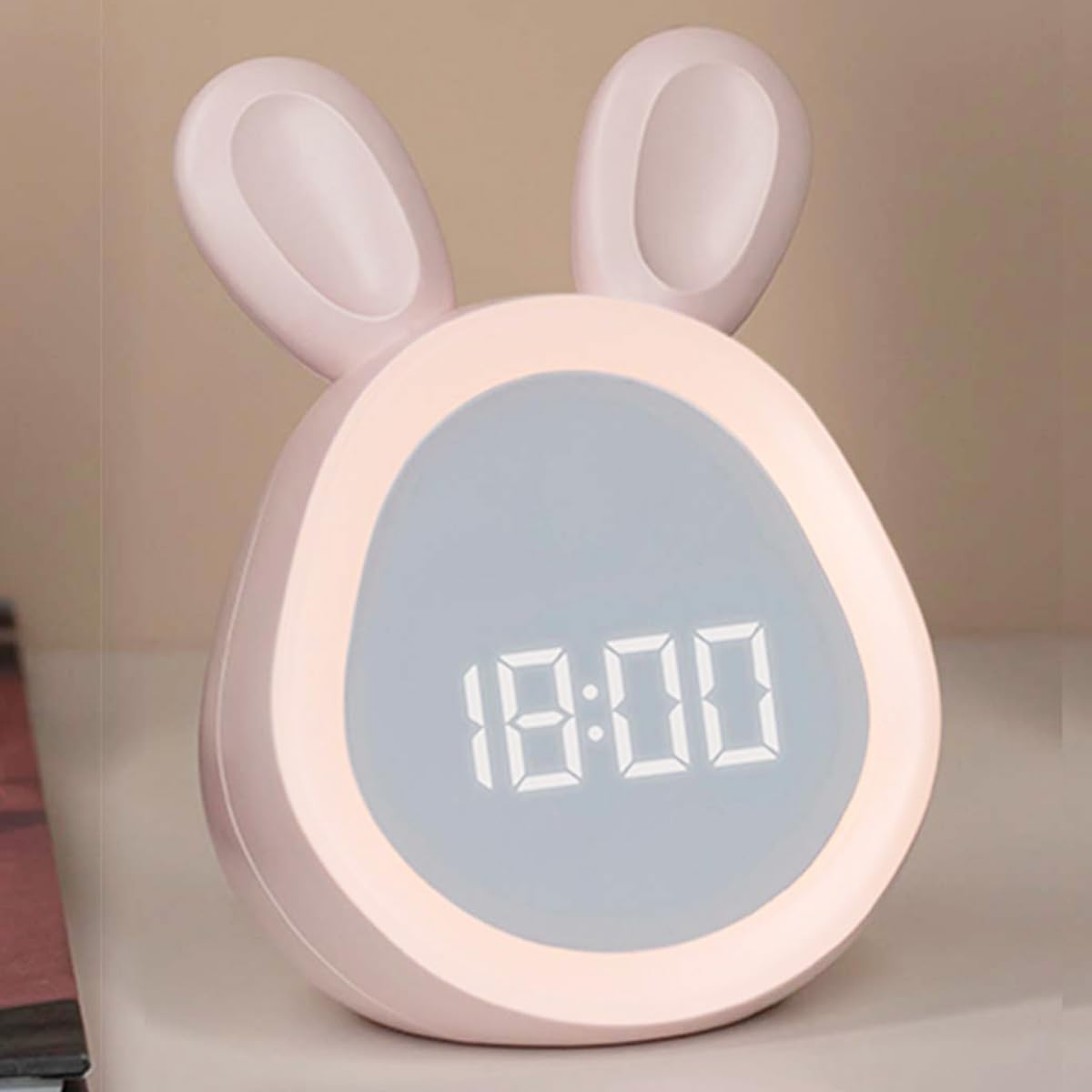Ein rosafarbener digitaler Wecker in Hasenform mit beleuchtetem Display zeigt 18:00 auf einem Tisch.