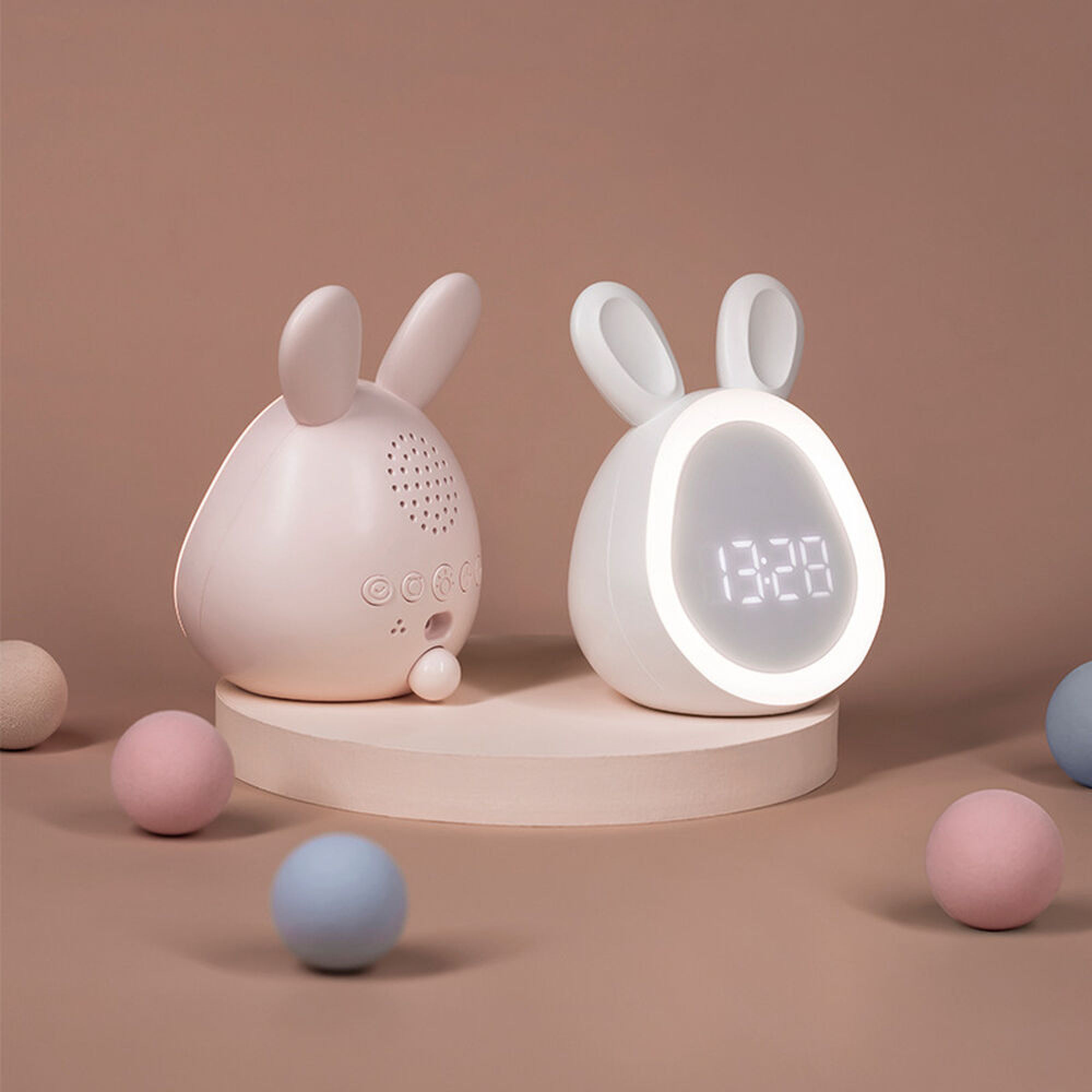 Zwei hasenförmige Wecker auf einem Podest, umgeben von kleinen pinken und blauen Bällen vor schlichtem Hintergrund.