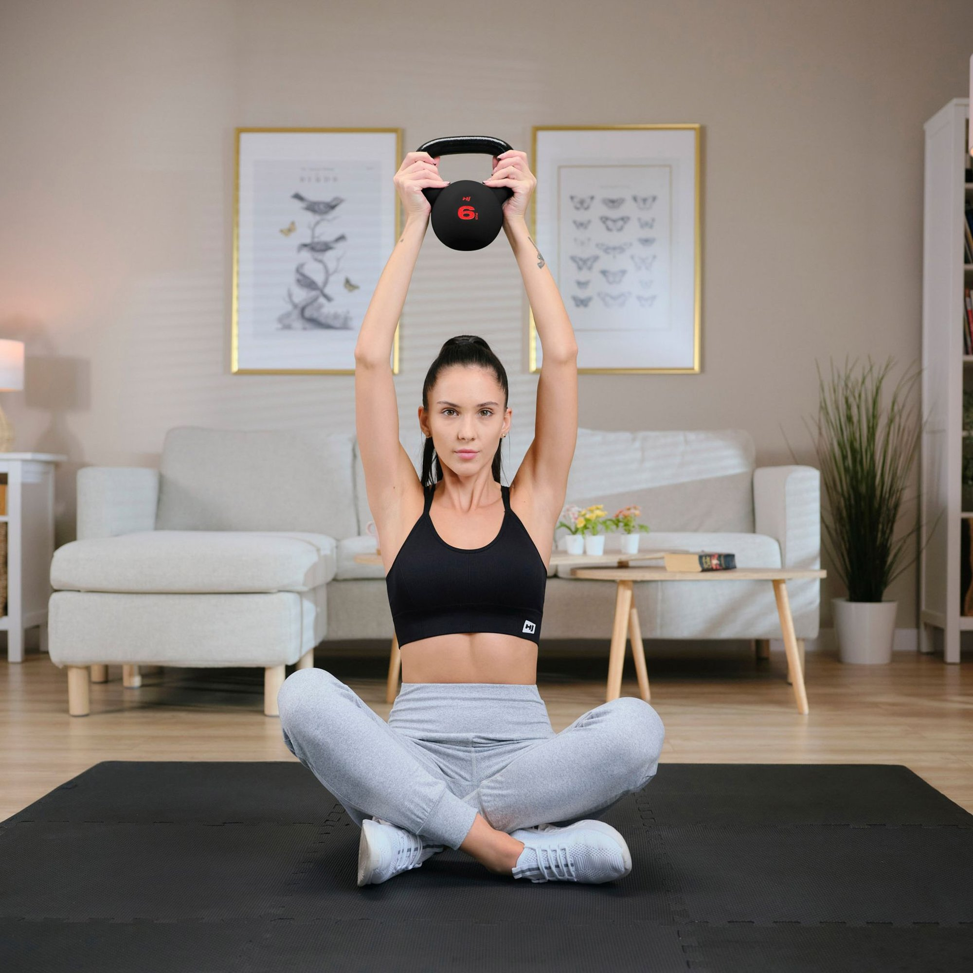 Eine Frau sitzt auf einer Matte im Wohnzimmer und hält einen Kettlebell über ihren Kopf.