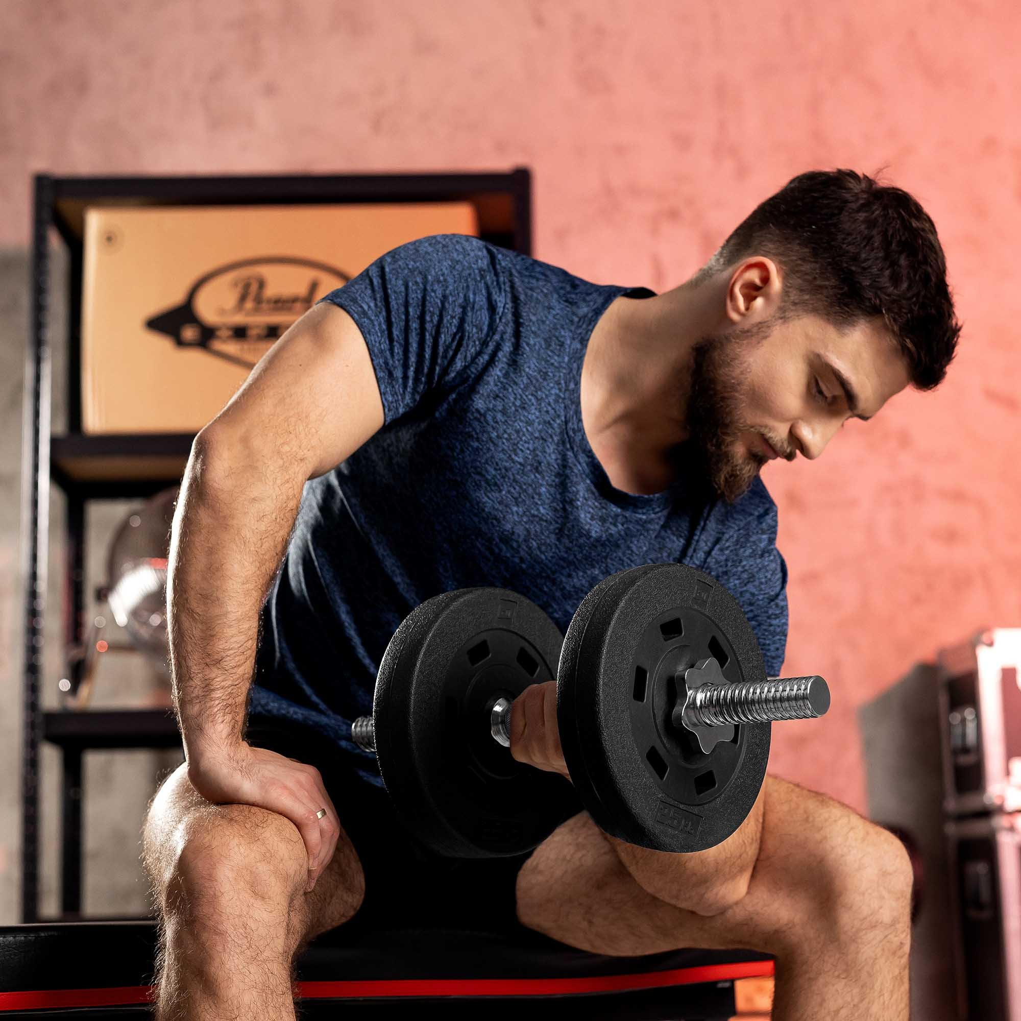 Ein bärtiger Mann in blauem Shirt sitzt auf einer Bank und hebt eine schwere Hantel in einem Fitnessraum.