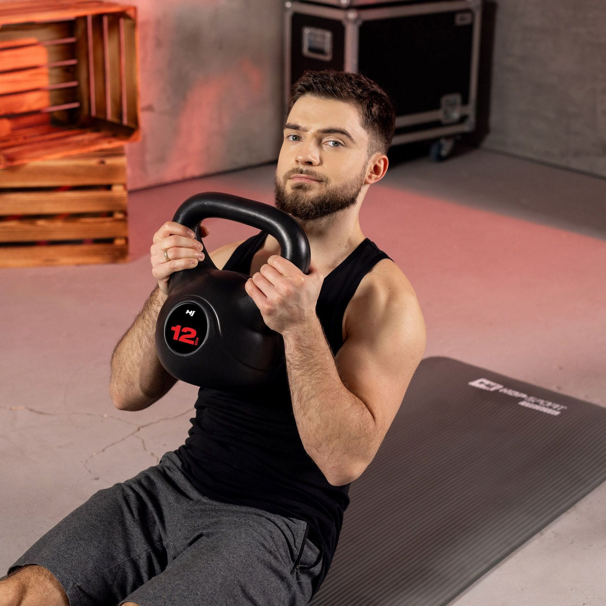 Ein Mann mit Bart sitzt auf einer Matte und hält eine 12 kg schwere Kettlebell vor seiner Brust.