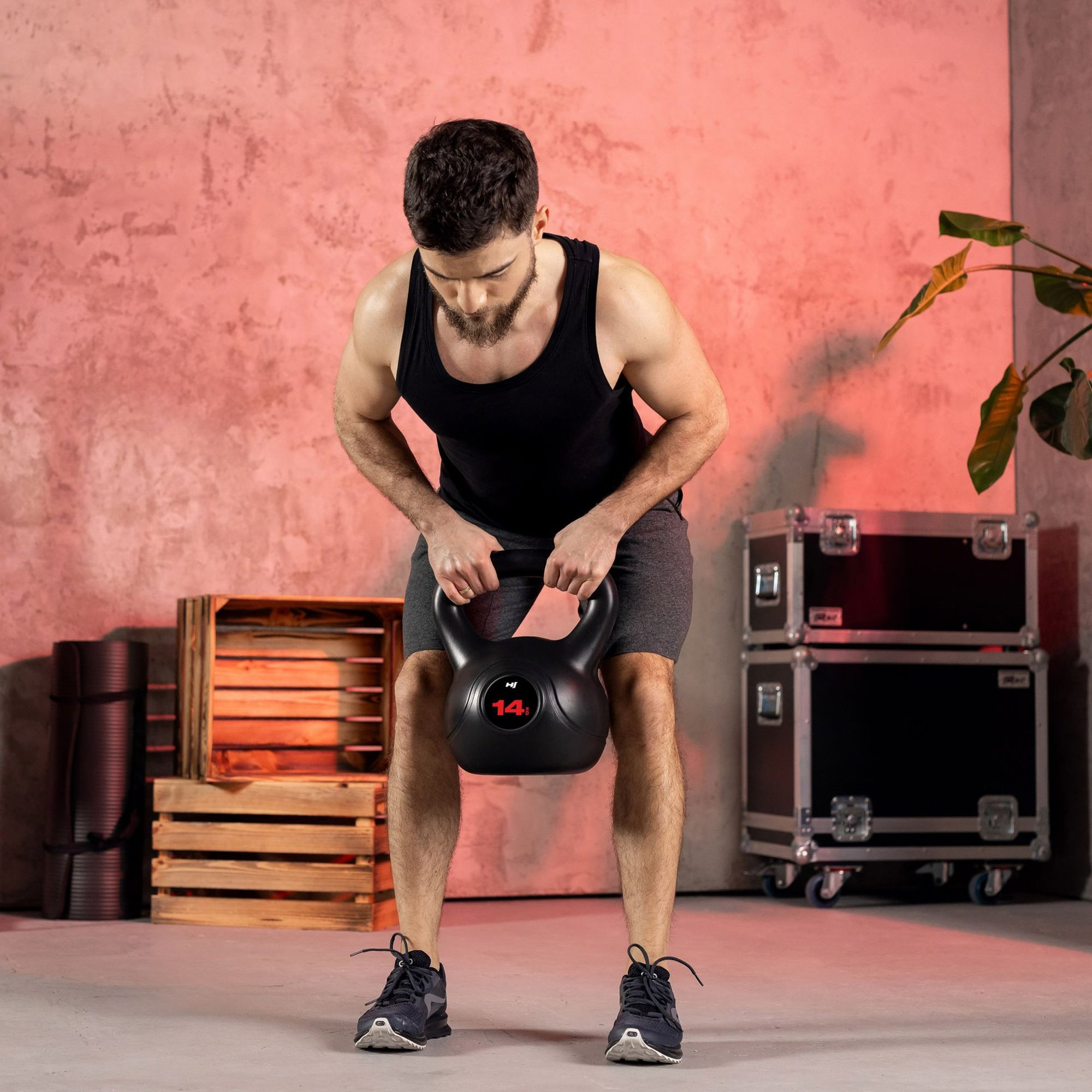 Ein Mann im Fitnessstudio hält eine schwarze Kugelhantel mit rotem 14kg-Etikett vor rosa Wand.