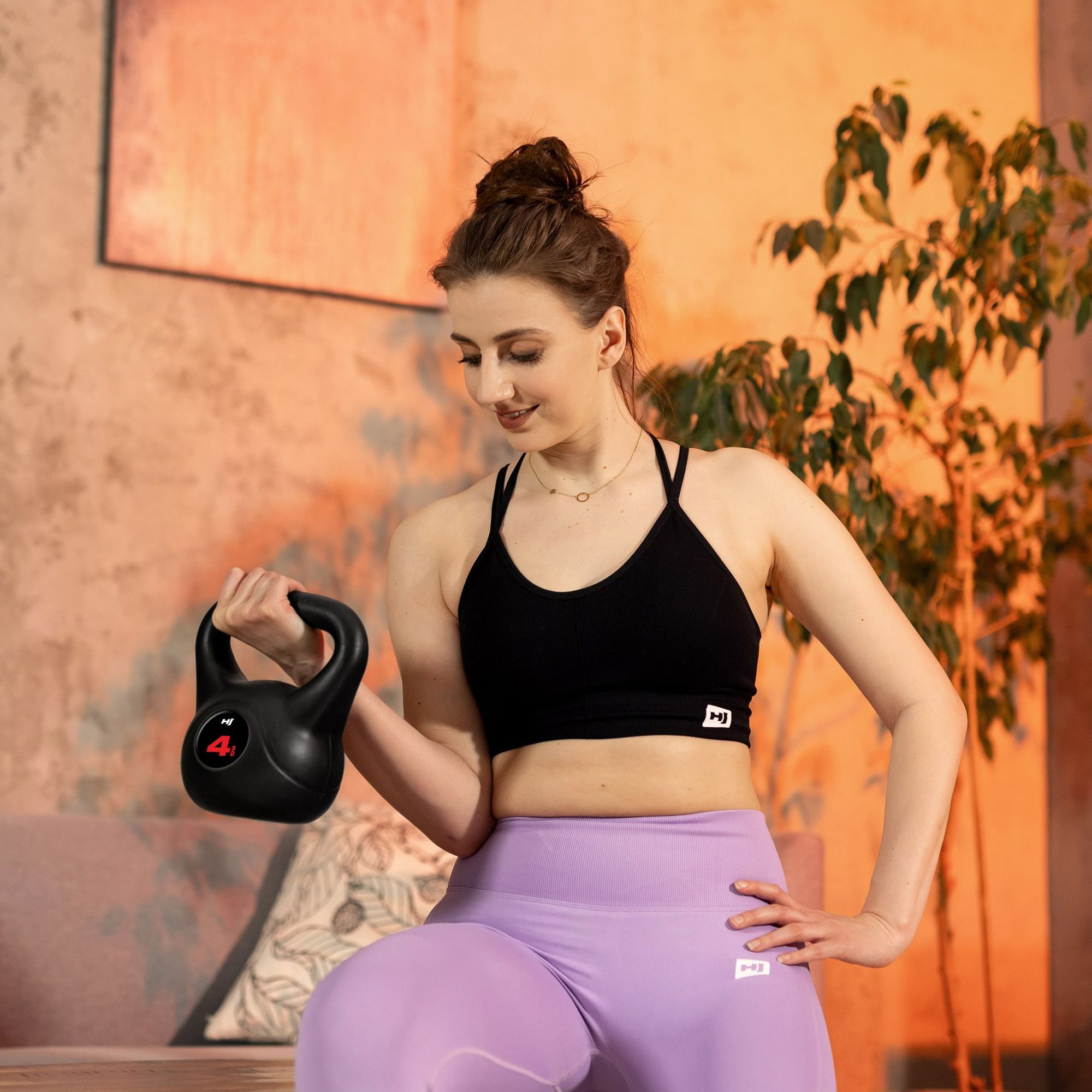 Eine Frau in Sportkleidung hebt eine schwarze 4kg-Kettlebell in einem gemütlichen Raum mit Pflanzen.