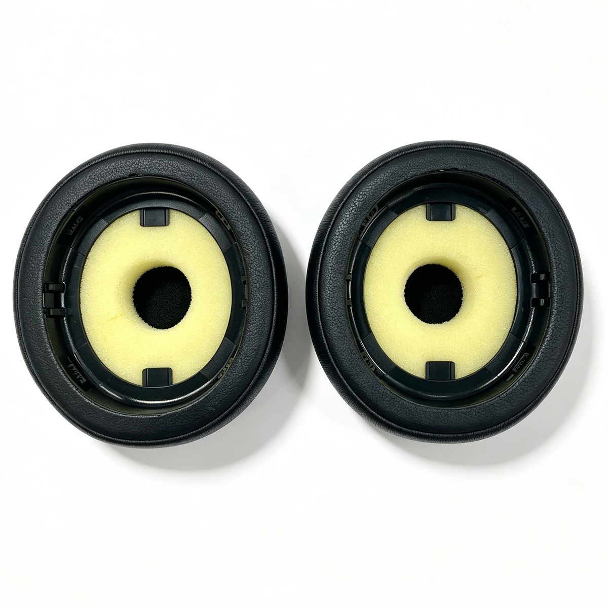 Two black circular headphone ear cushions with yellow foam interiors, viewed from the back on a white background.