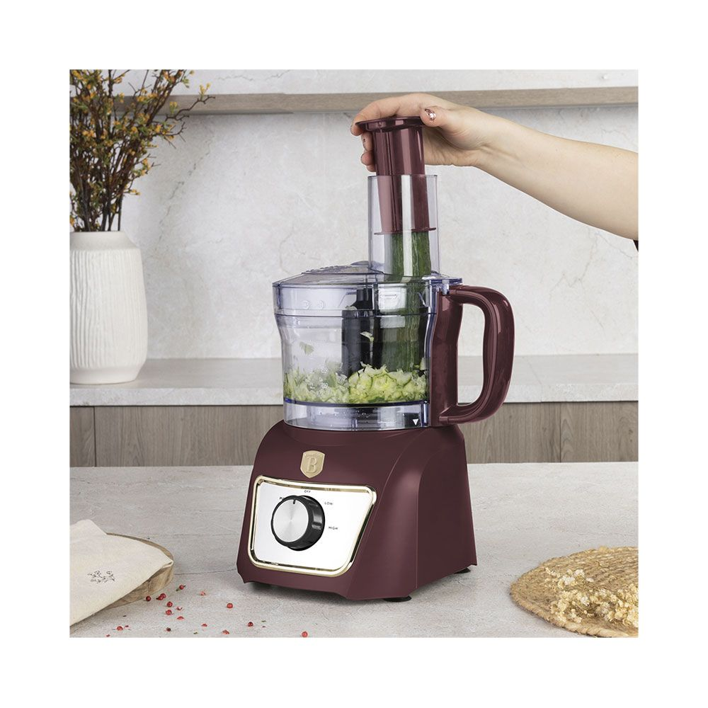 Une main utilise un robot culinaire rouge foncé pour hacher un concombre sur un comptoir.