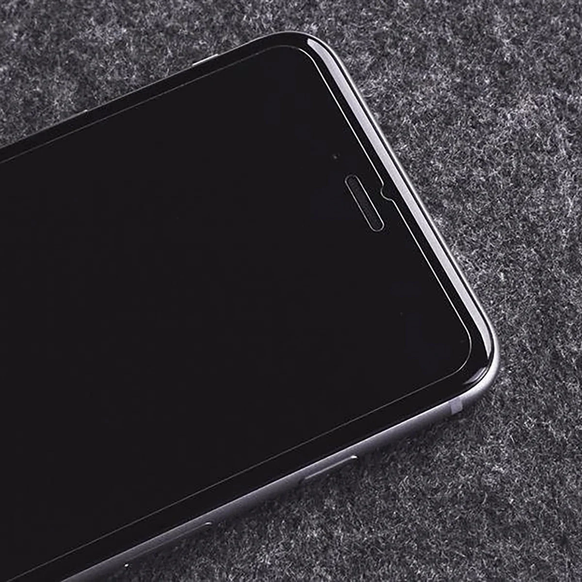 Primo piano di uno smartphone nero con pellicola protettiva su una superficie scura testurizzata.
