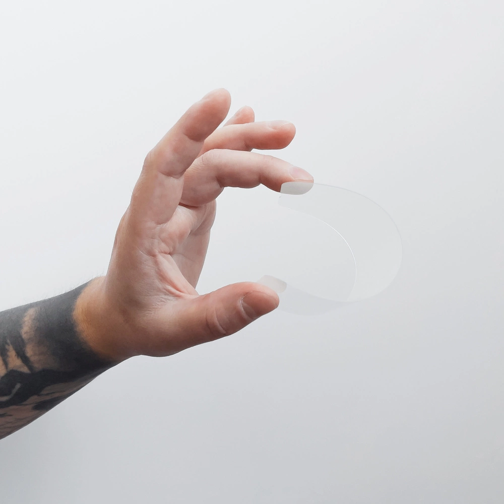 Una mano con un tatuaggio scuro tiene una striscia di plastica trasparente curva su sfondo bianco.
