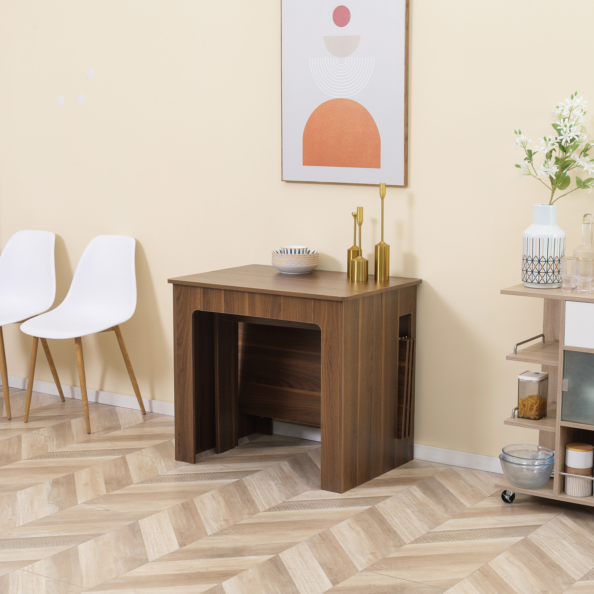 Una mesa de madera en una habitación beis con dos sillas blancas, arte en la pared, candelabros dorados y una estantería.