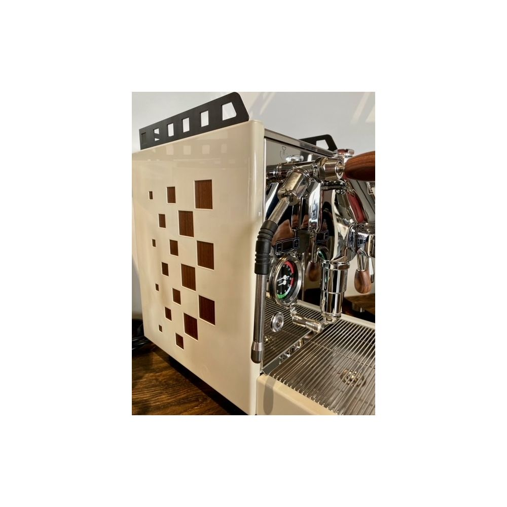 Una macchina da caffè espresso bianco crema con dettagli in legno, parti cromate e motivo quadrato sul lato si trova su un bancone.