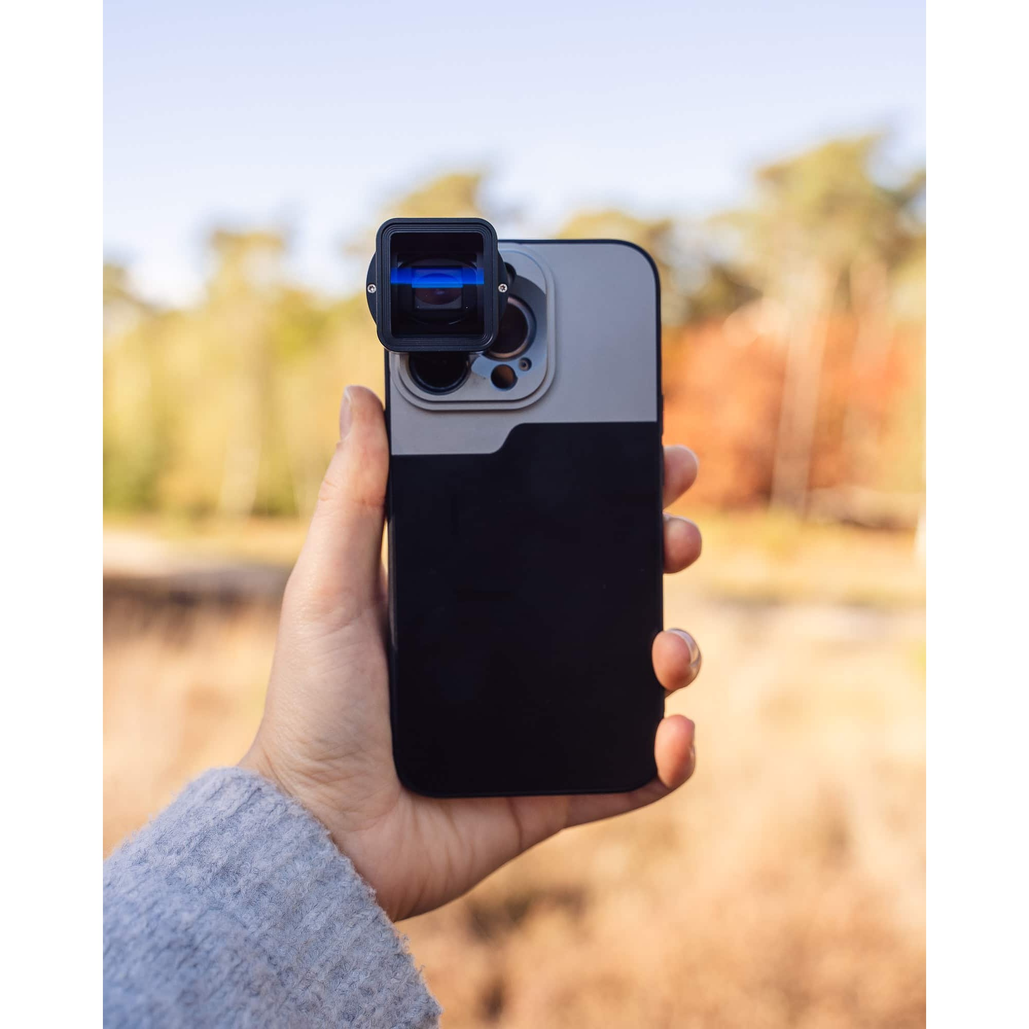 Eine Hand hält ein Smartphone mit einem aufgesetzten quadratischen Anamorphot-Objektiv vor einer verschwommenen Waldkulisse.