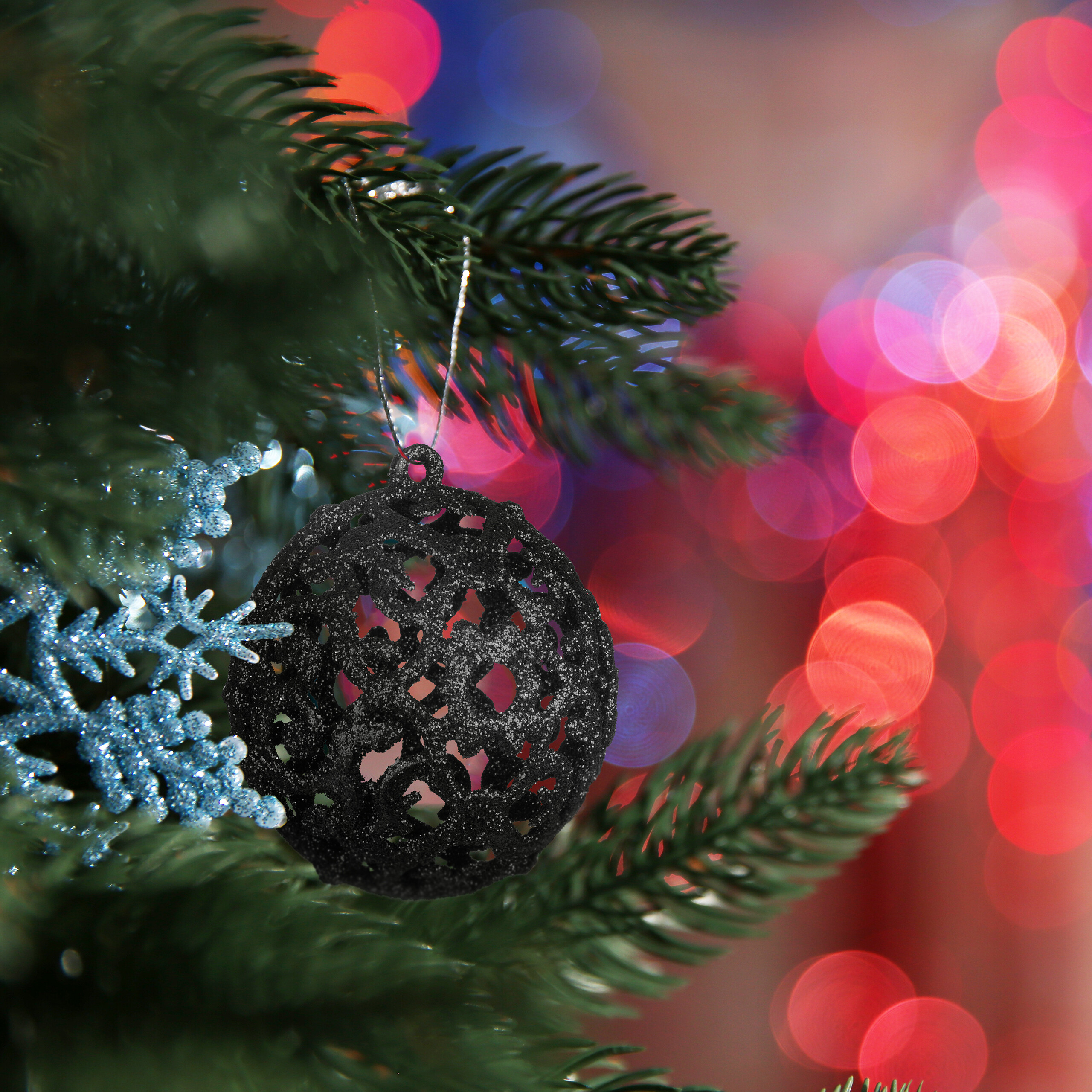 Eine schwarze, glitzernde Weihnachtskugel und blaue Schneeflocken hängen an einem Tannenbaum vor verschwommenem Hintergrund.