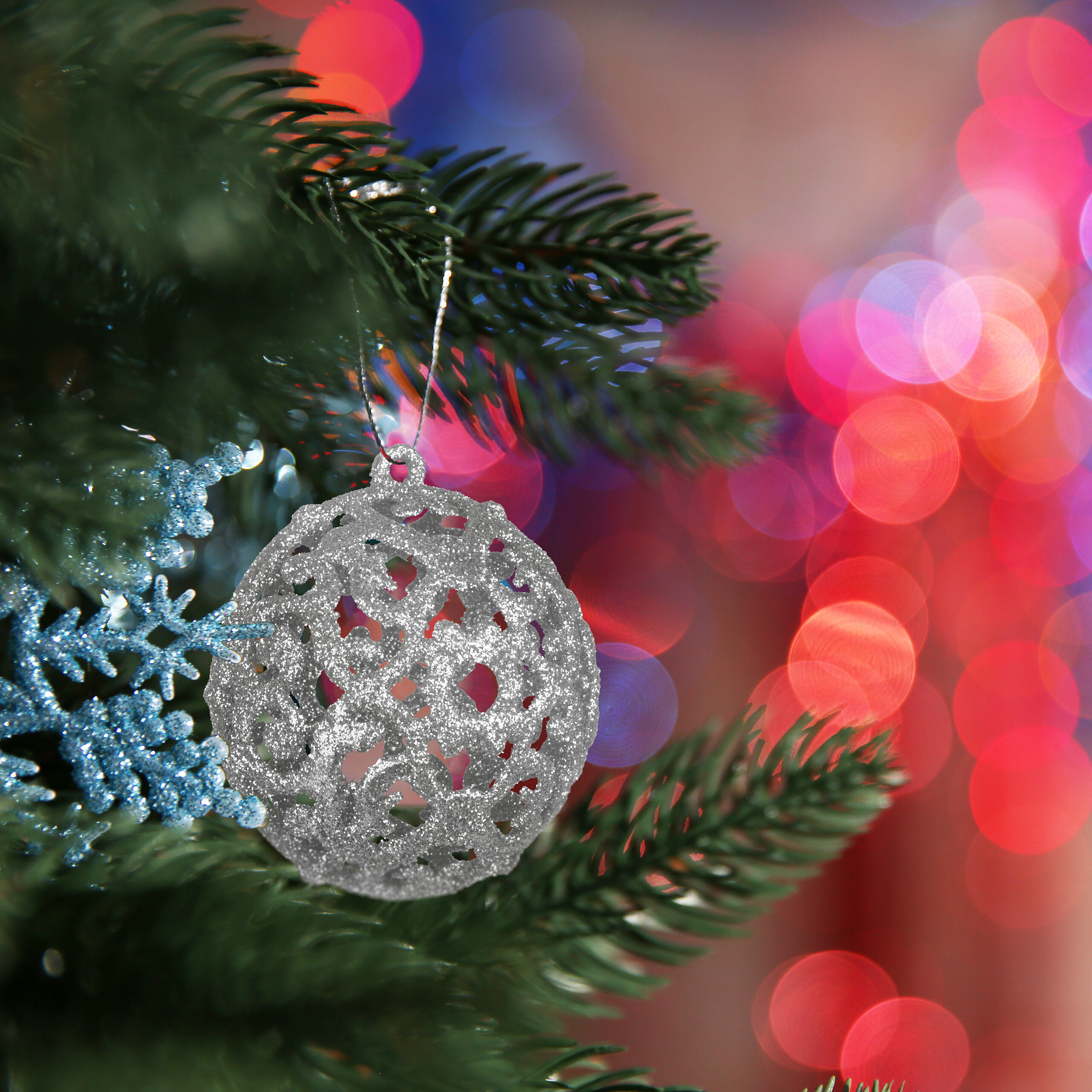 Eine glitzernde silberne Weihnachtskugel hängt an einem grünen Zweig vor bunten Lichtern.