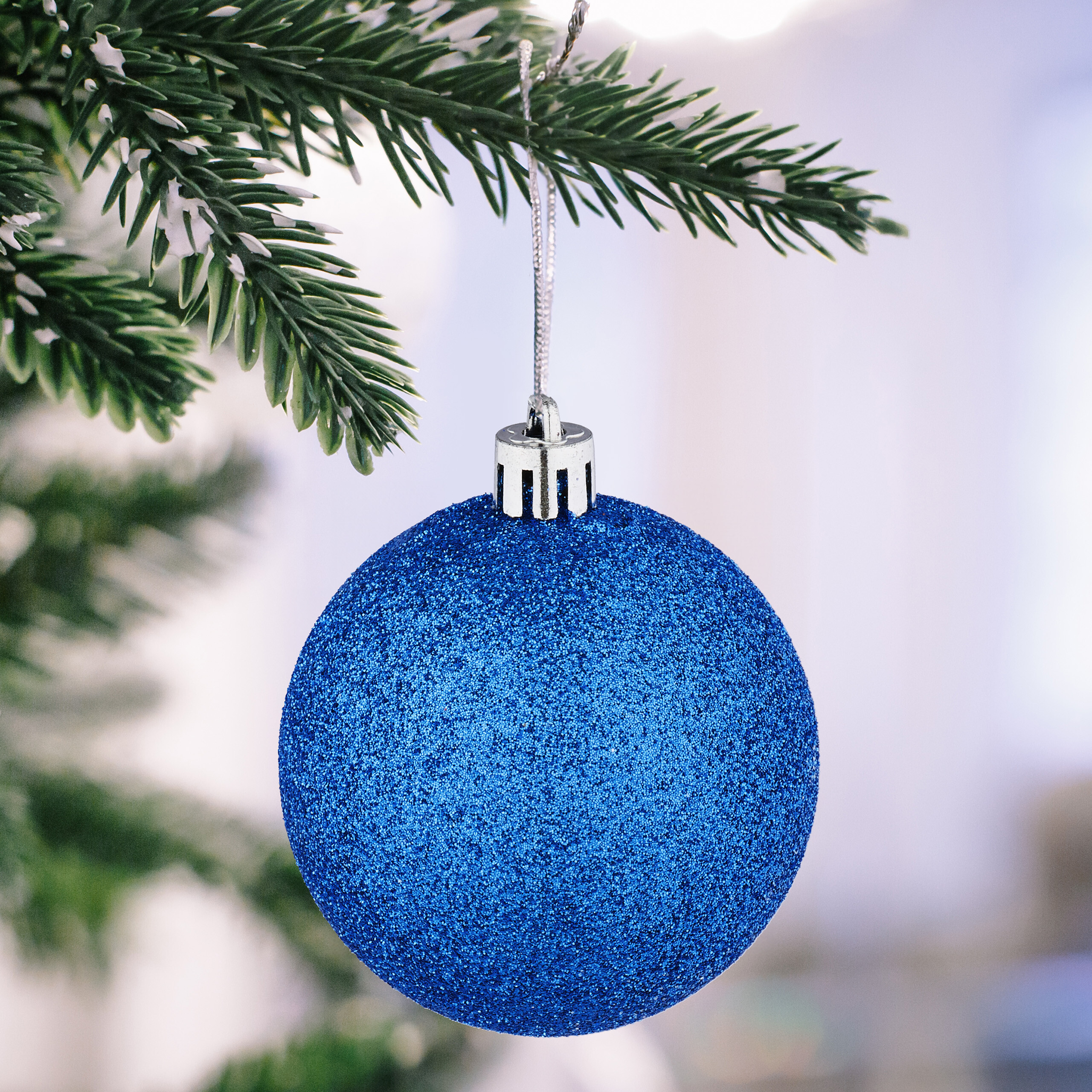 Eine glitzernde blaue Weihnachtskugel hängt an einem verschneiten Tannenzweig.