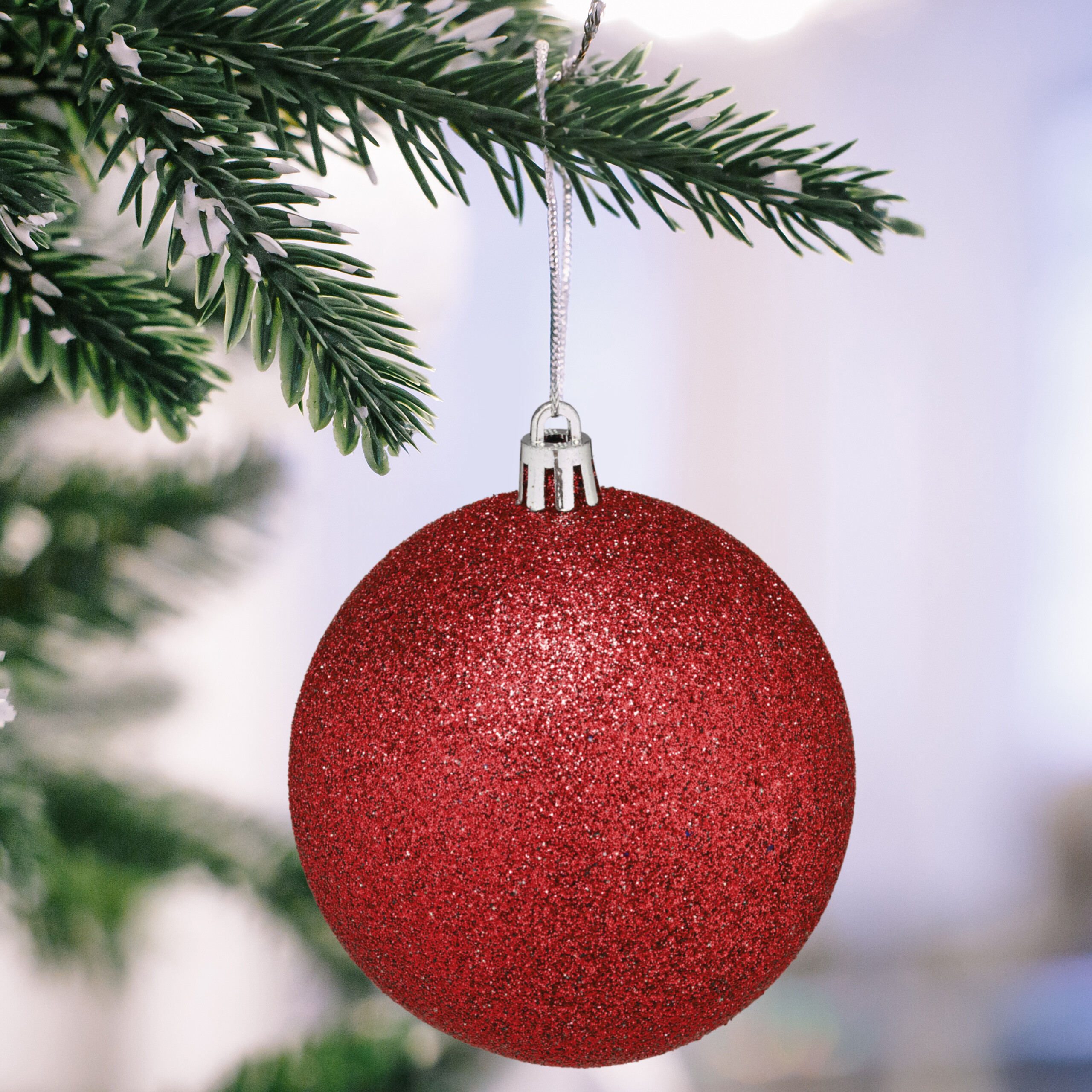 Eine glitzernde rote Weihnachtskugel hängt an einem frostigen Tannenzweig vor verschwommenem Hintergrund.