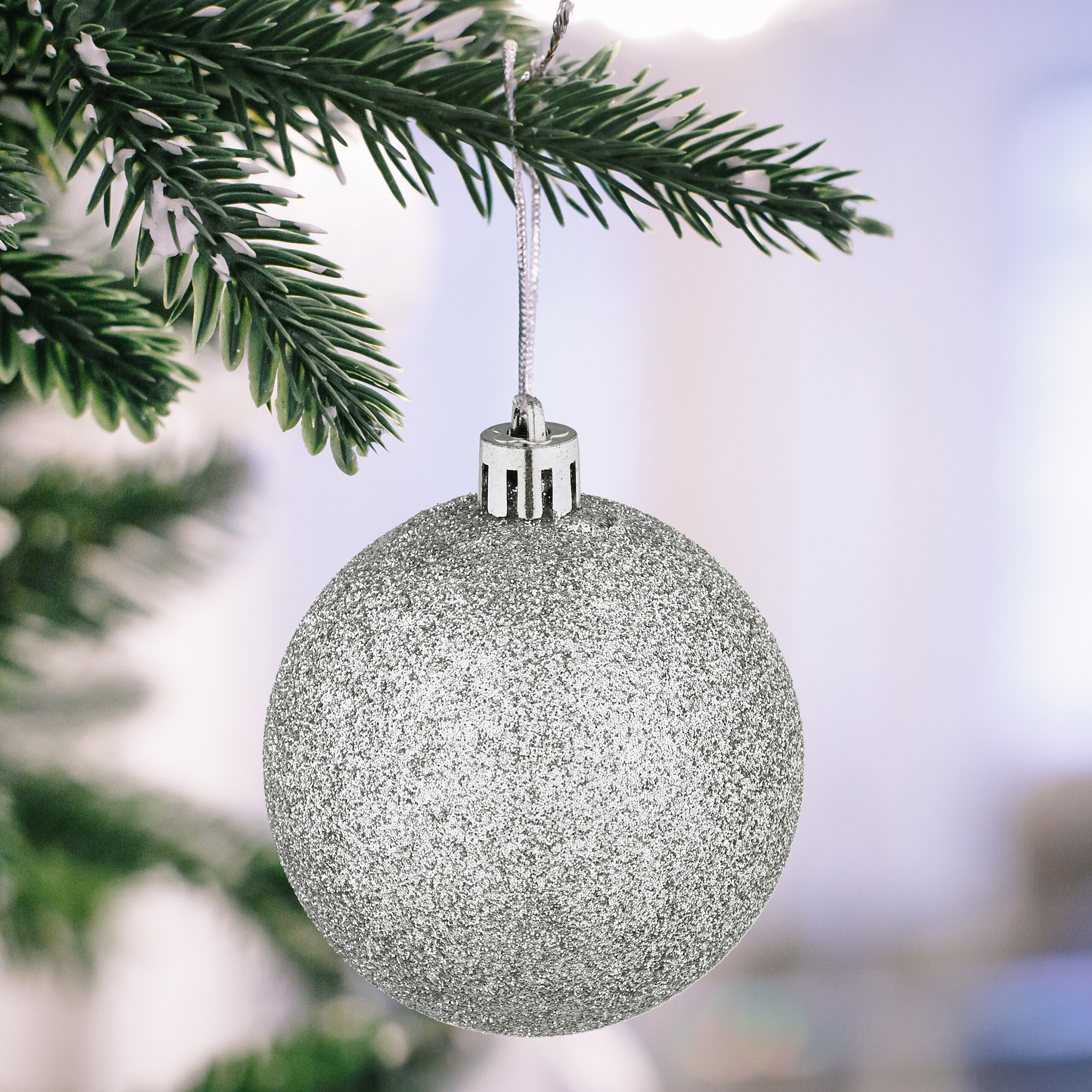 Eine glitzernde silberne Christbaumkugel hängt an einem schneebedeckten Tannenzweig vor verschwommenem Hintergrund.