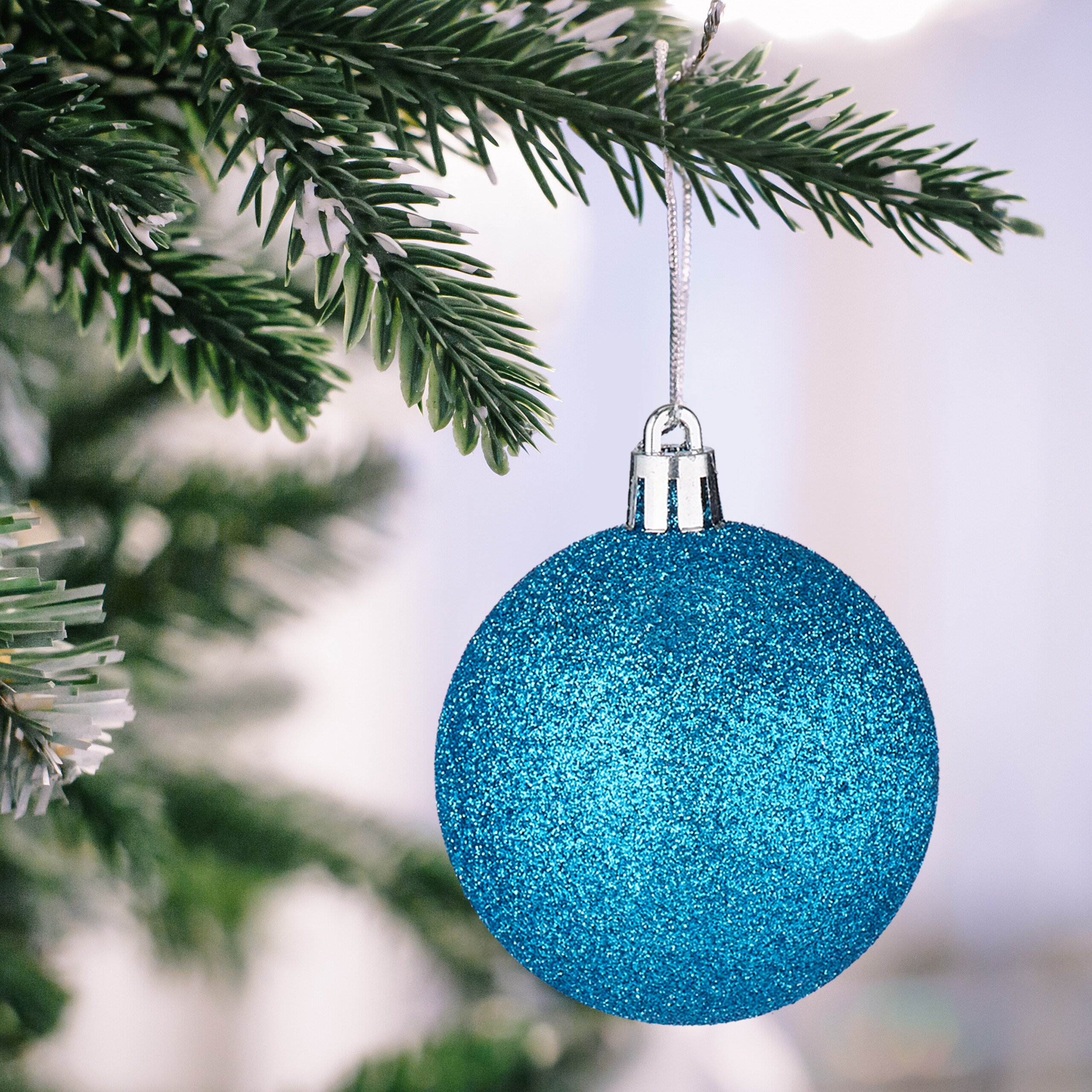 Eine glitzernde blaue Weihnachtskugel hängt an einem schneebedeckten Tannenzweig.