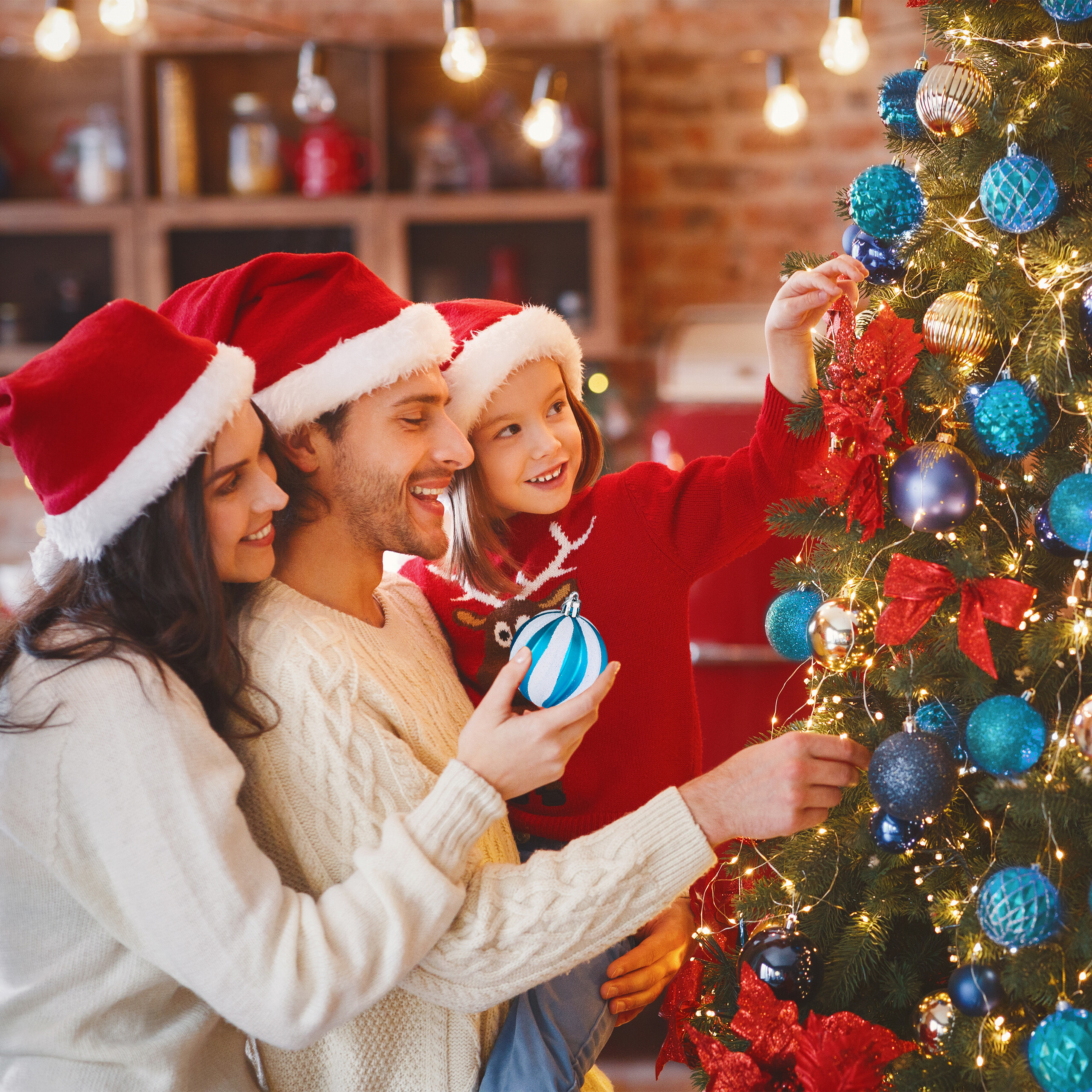 Eine Familie mit Weihnachtsmützen dekoriert zusammen einen Weihnachtsbaum mit blauen und goldenen Kugeln.