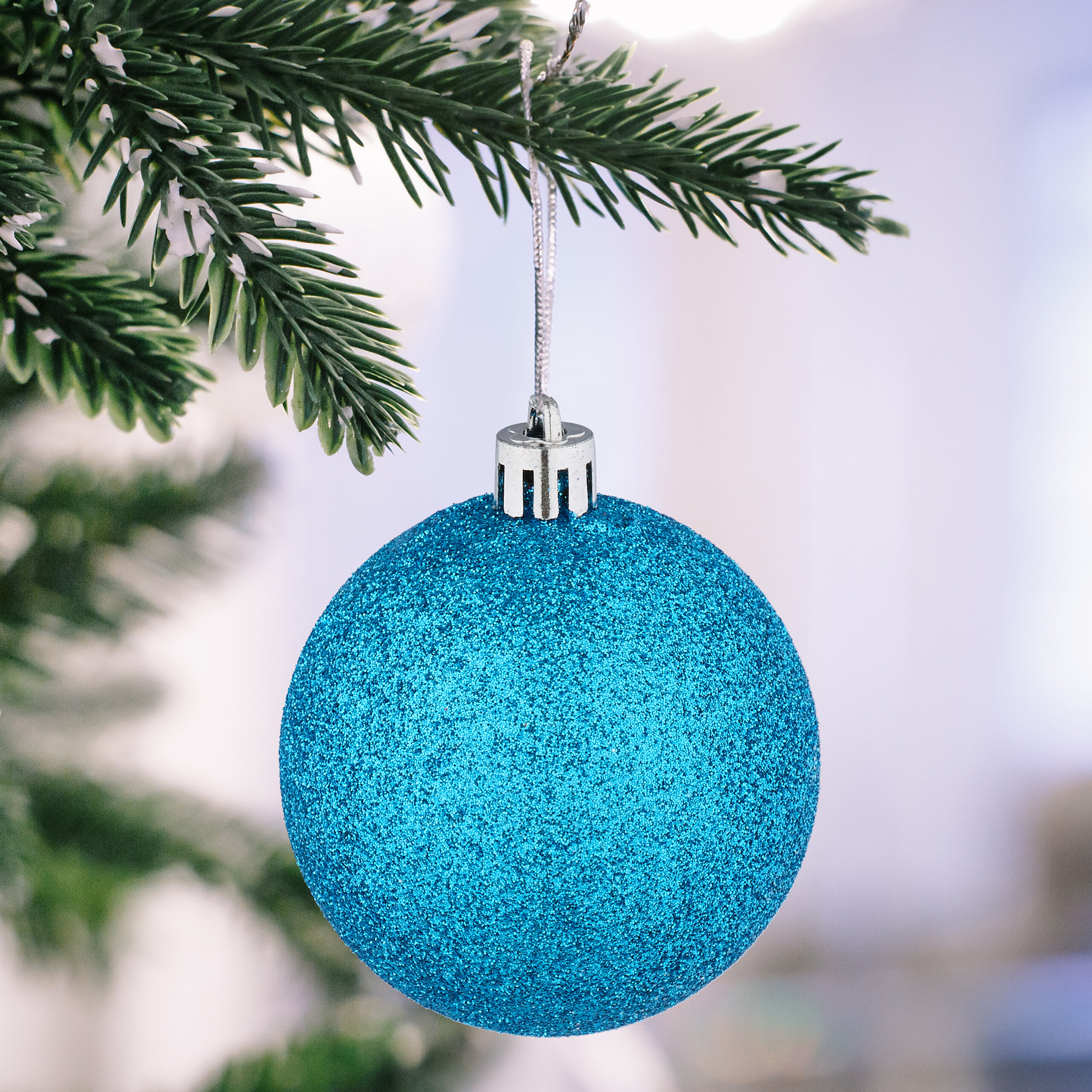 Eine glitzernde blaue Weihnachtskugel hängt an einem verschneiten Tannenzweig.