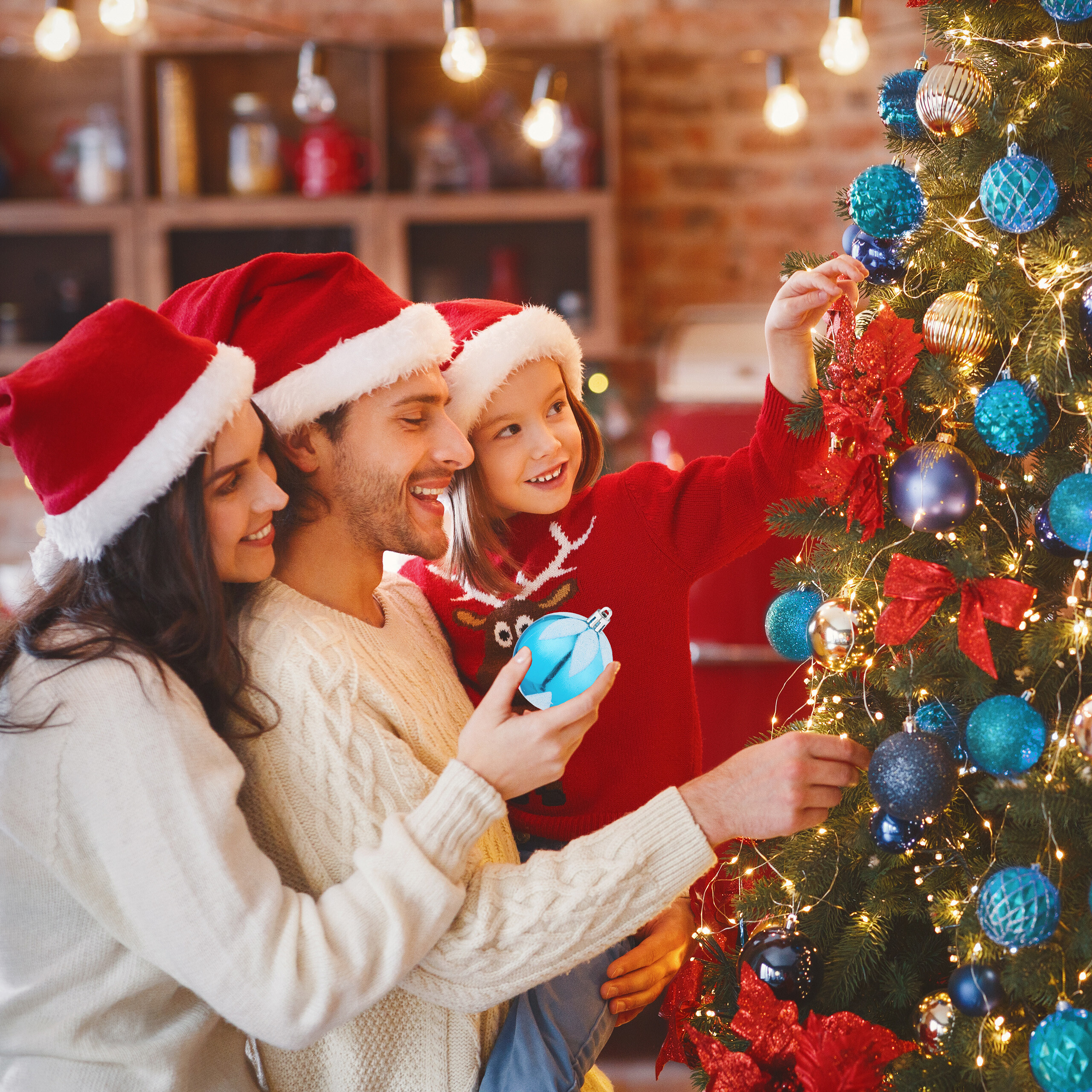 Familie mit Weihnachtsmützen dekoriert gemeinsam einen Weihnachtsbaum in einem warm erleuchteten Zimmer.