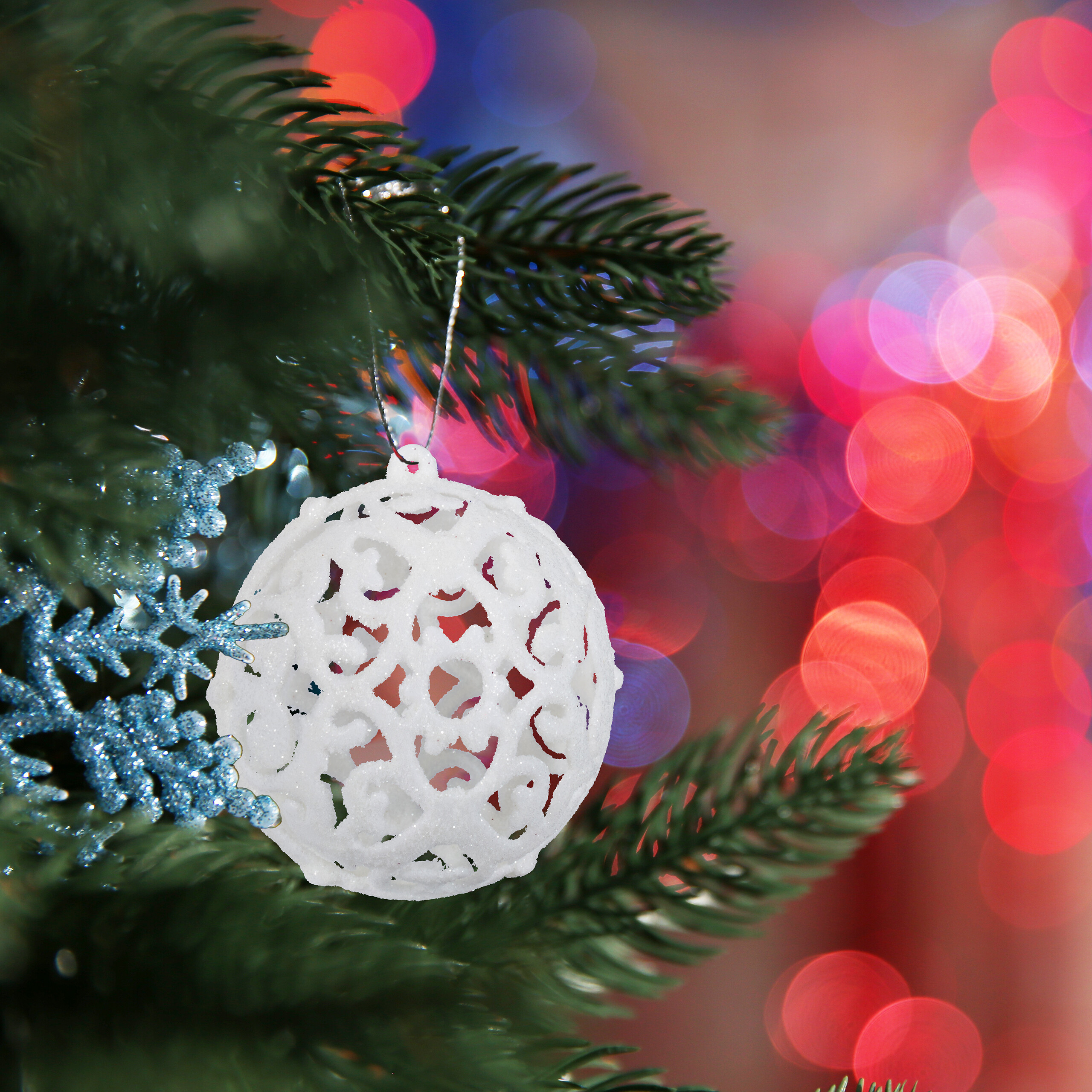 Eine weiße Weihnachtskugel hängt an einem grünen Zweig vor verschwommenem, bunten Hintergrund.