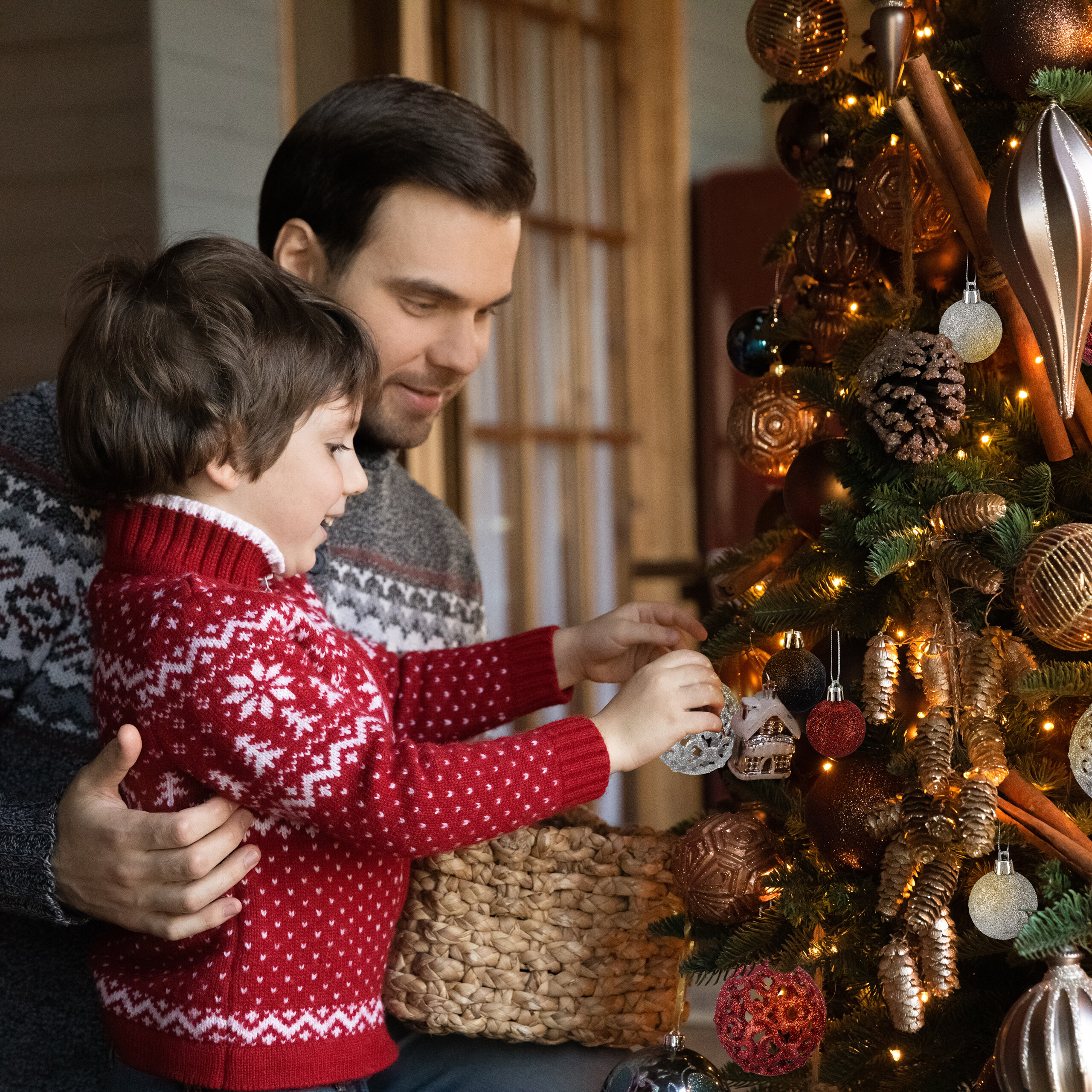 Ein Vater und sein kleiner Sohn in Weihnachtspullovern dekorieren einen funkelnden Weihnachtsbaum.