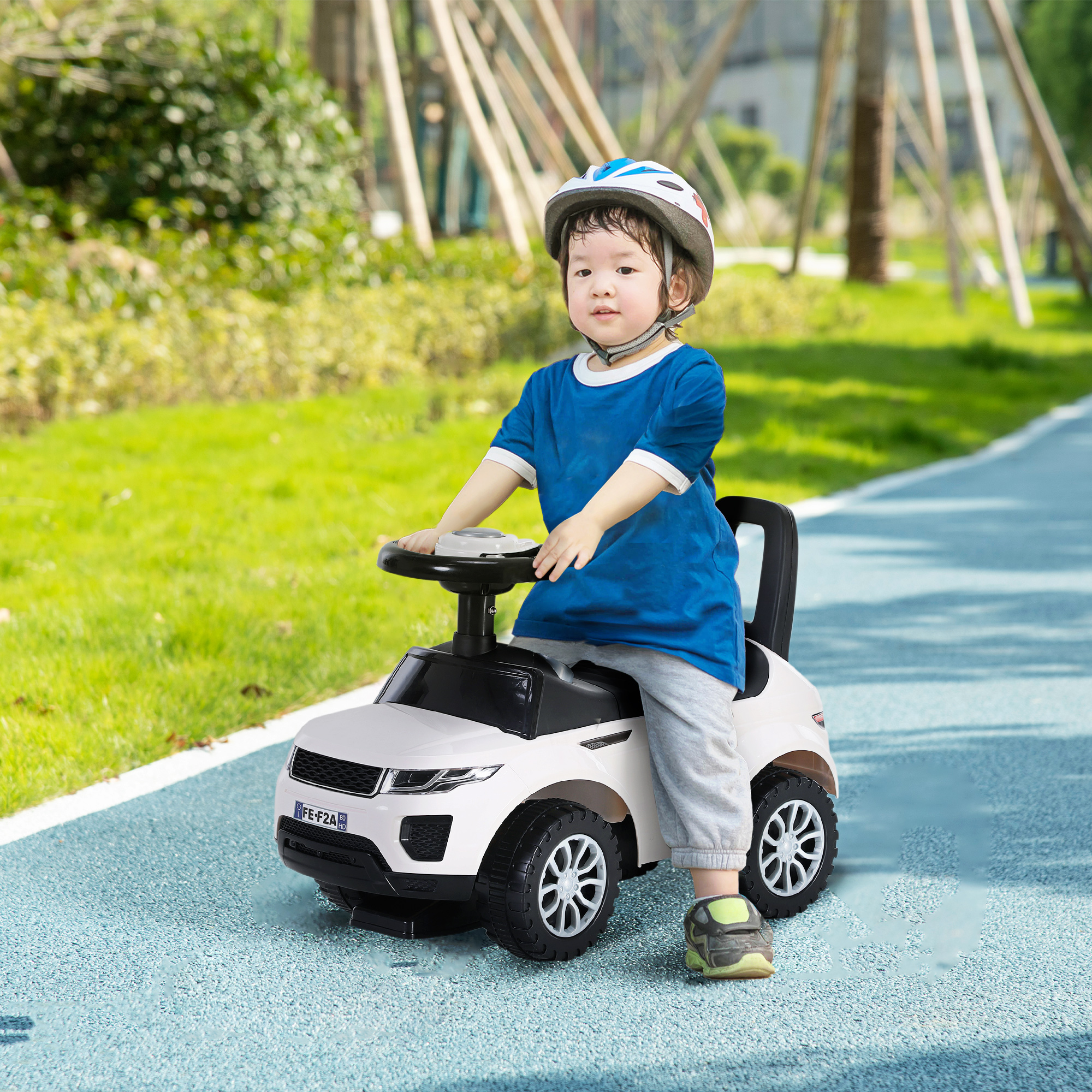 Un bambino con camicia blu e casco guida un'auto giocattolo bianca in un parco.