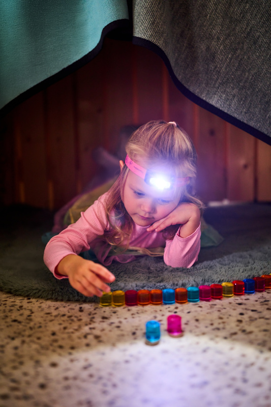 Ein blondes Mädchen mit Stirnlampe liegt in einer Deckenhöhle und spielt mit bunten Glasperlen auf dem Boden.