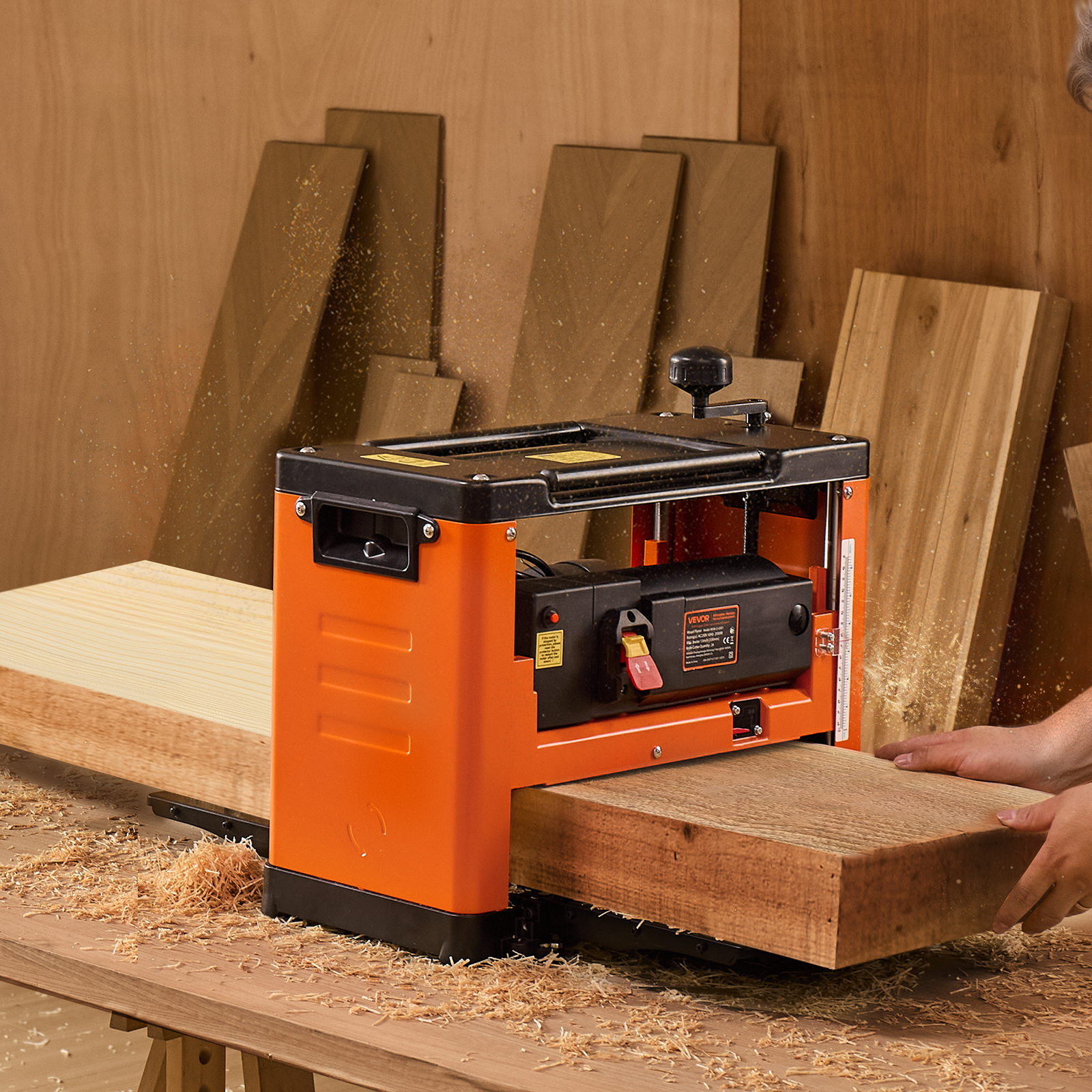 Una regruesadora naranja cepilla una tabla de madera, rodeada de virutas y tablones en el fondo.
