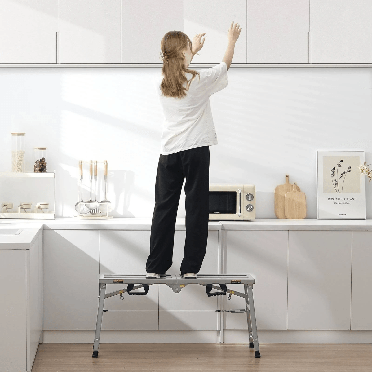 Una mujer de pie en un taburete de plataforma en una cocina blanca alcanza un armario.