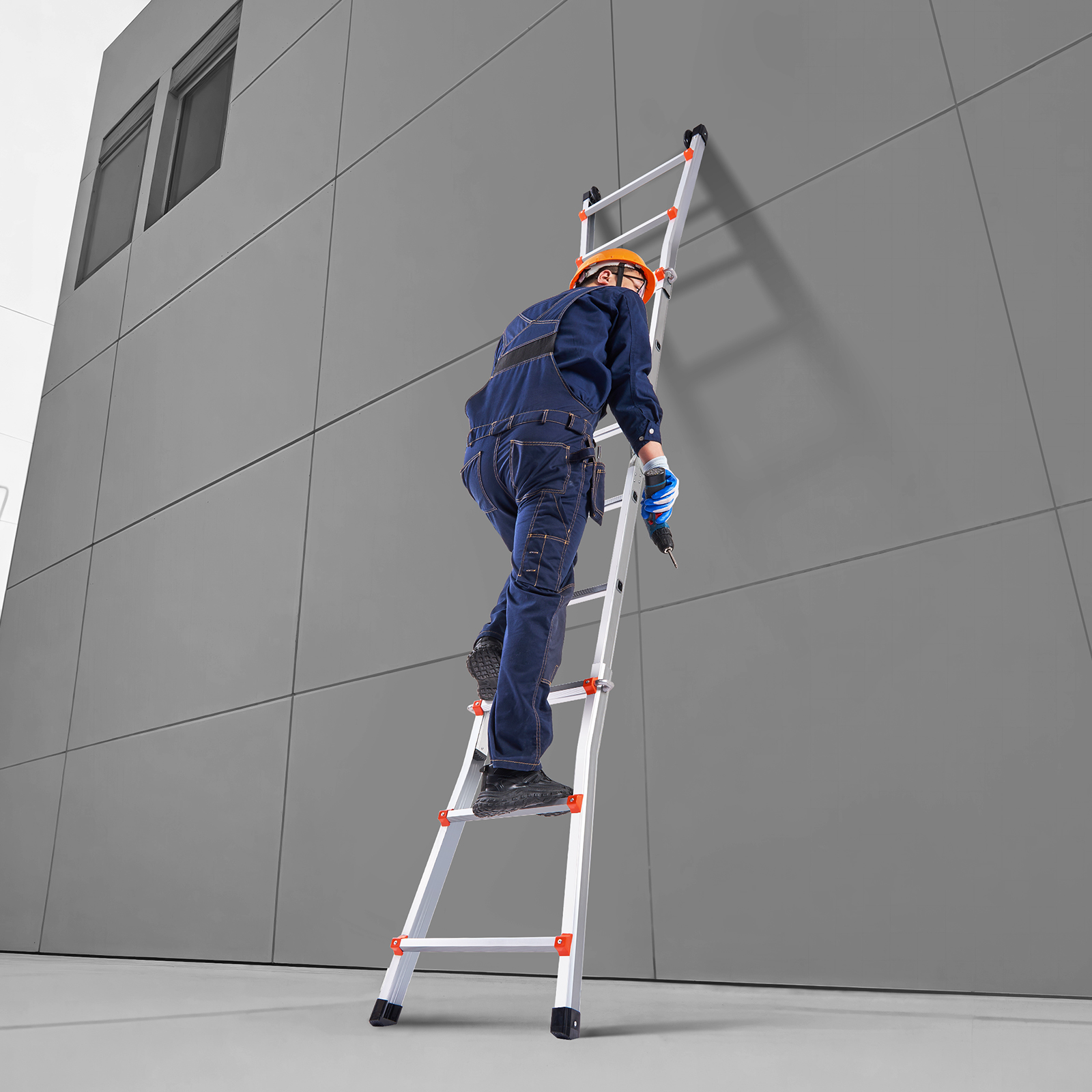 Un trabajador con mono azul y casco sube por una escalera metálica contra una pared gris.