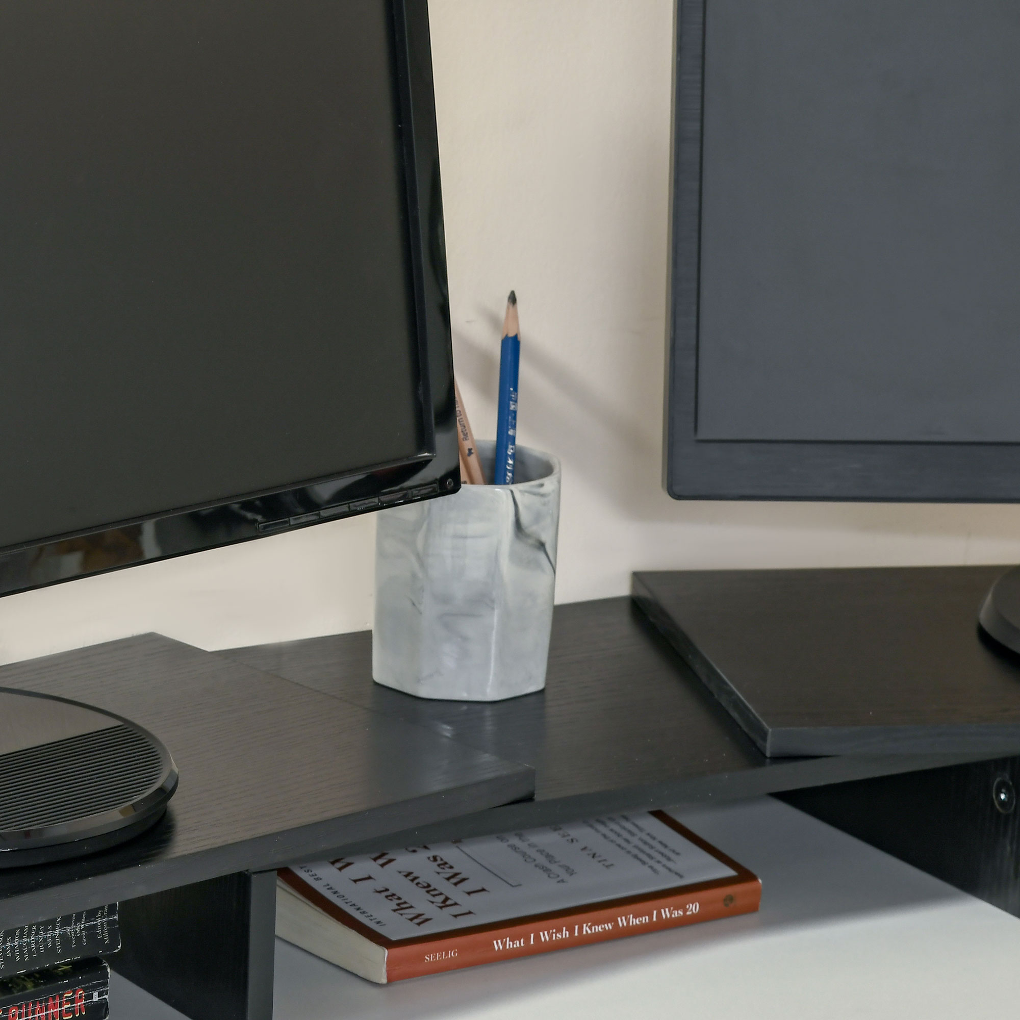 Dos monitores en un soporte de escritorio negro, un portalápices entre ellos y libros debajo.