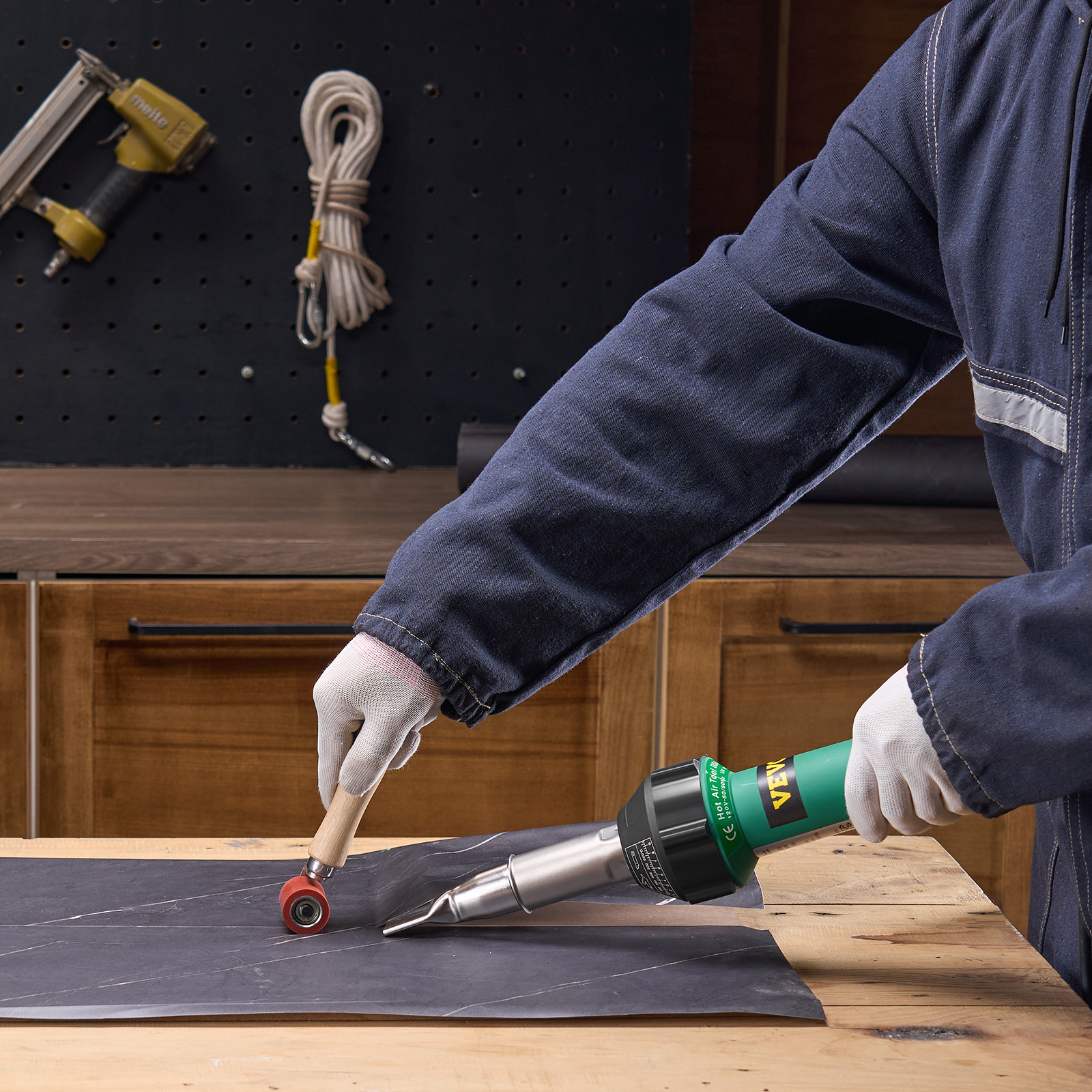 Persona con ropa de trabajo azul y guantes usa pistola de calor y rodillo sobre material oscuro en mesa de madera.