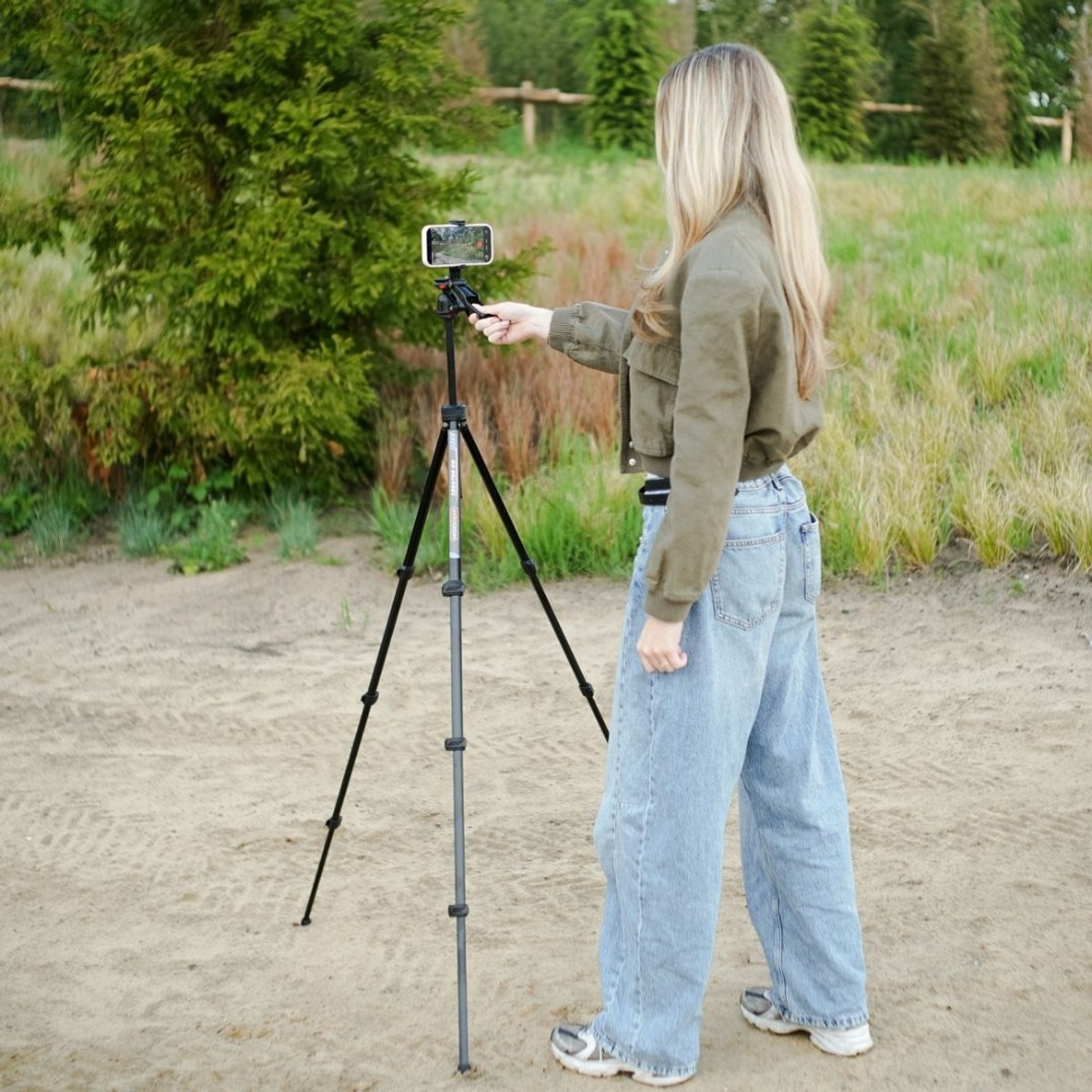 A blonde woman in an olive jacket and jeans films with a smartphone on a tripod outdoors.