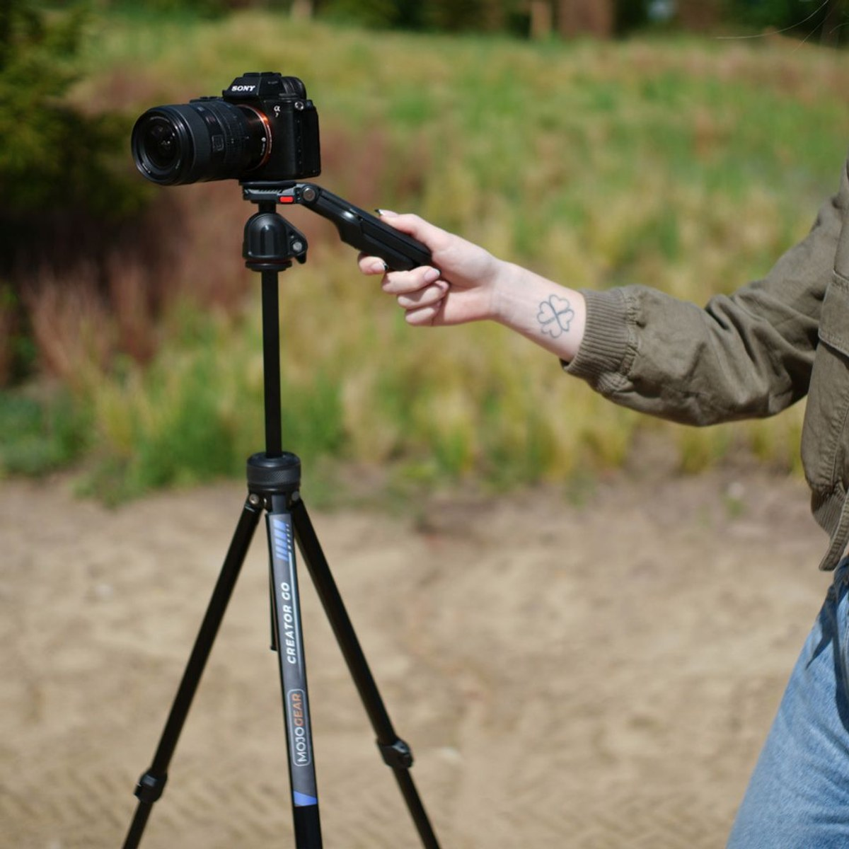 A person holds a black camera mounted on a tripod outdoors with green foliage and sandy ground in the background.