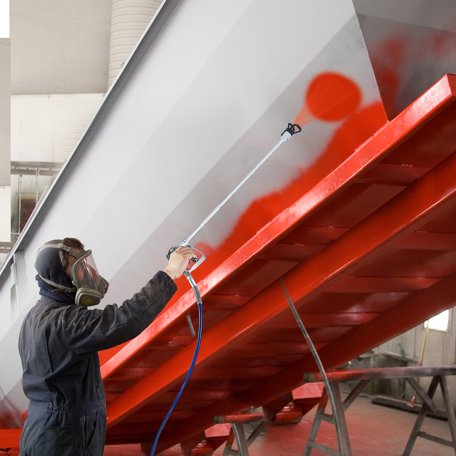 Un trabajador con mascarilla protectora rocía pintura roja en una gran estructura de metal gris en un taller.