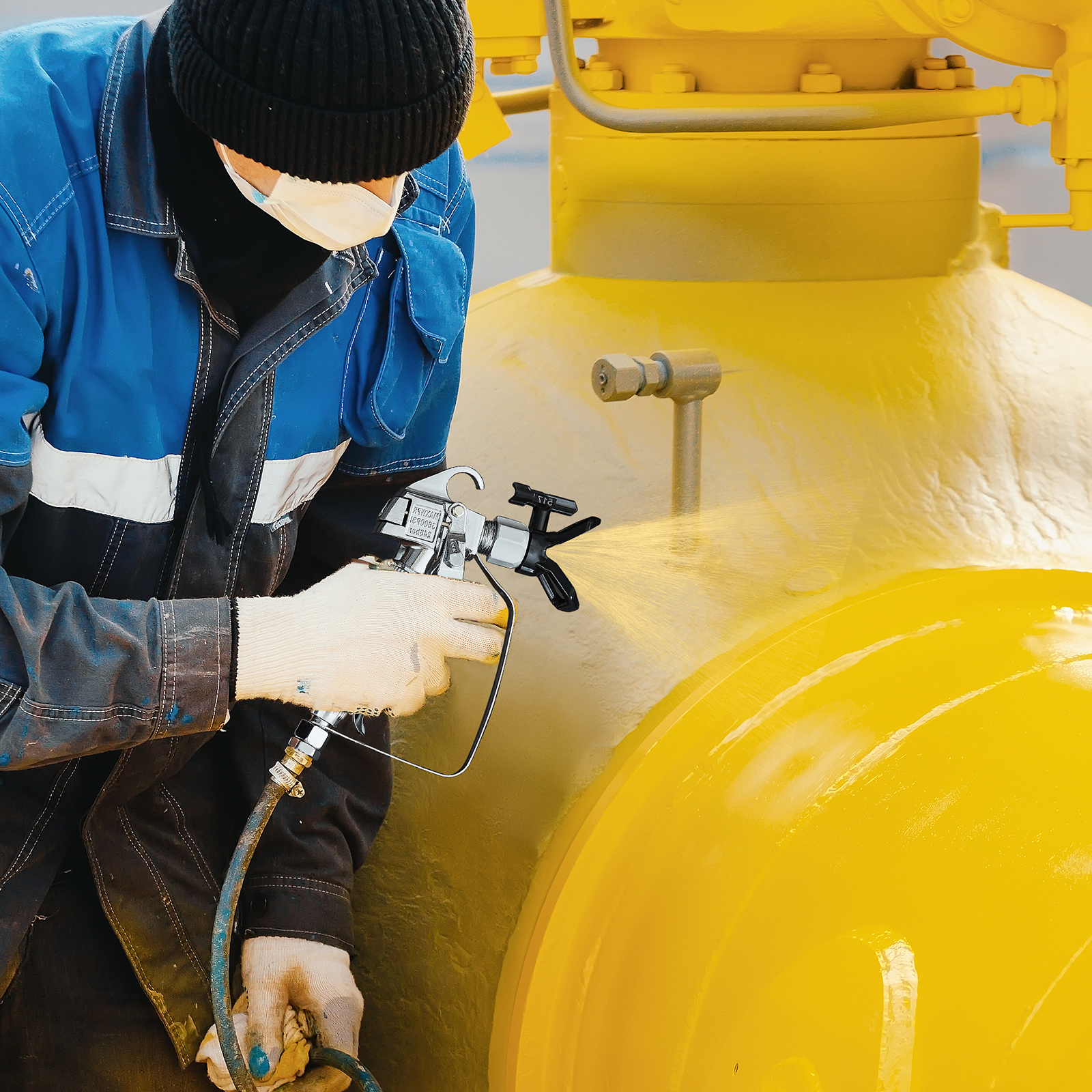 Un trabajador con equipo de protección azul pinta una gran máquina industrial amarilla.