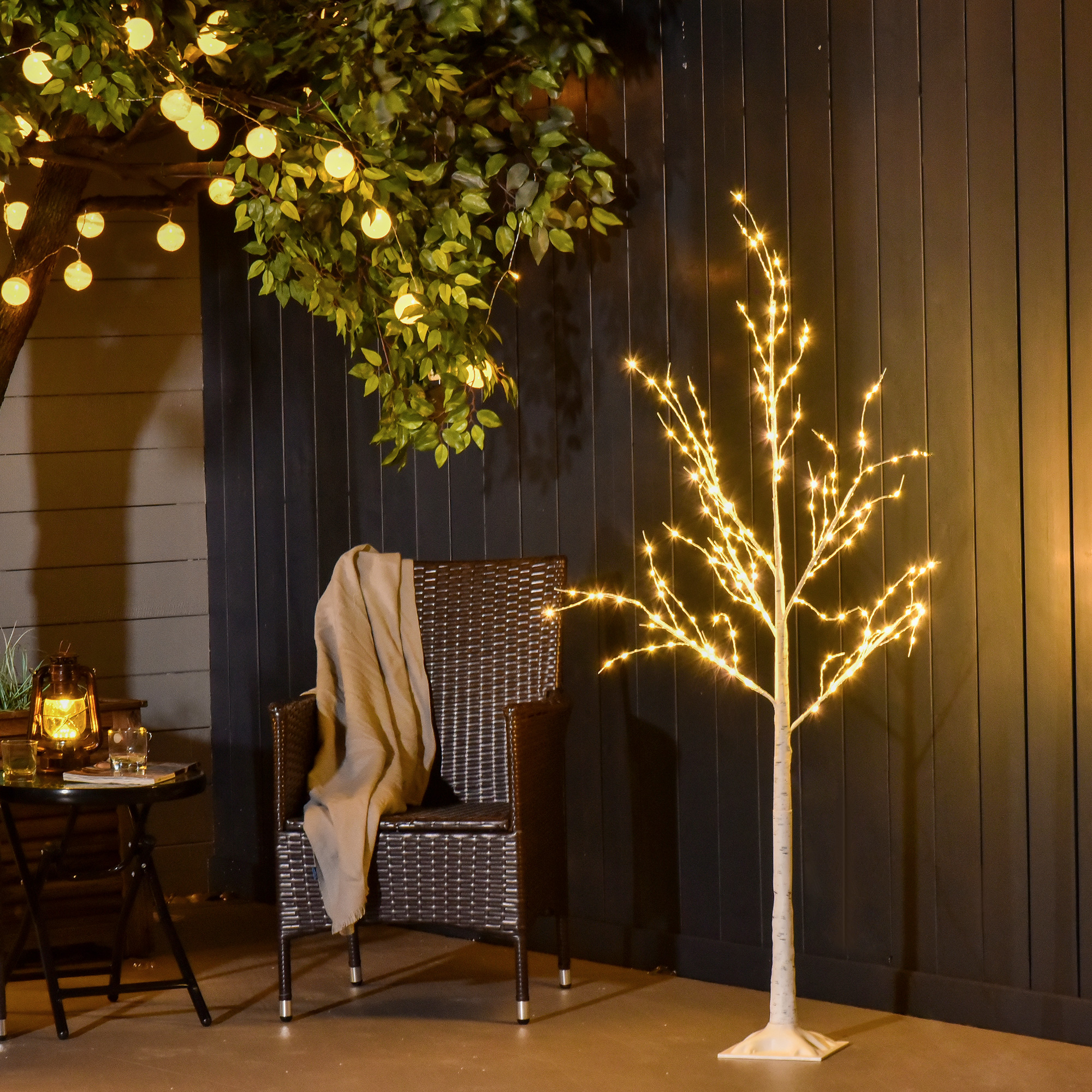 Árbol artificial iluminado, sillón de mimbre con manta y mesa pequeña con linterna contra una pared oscura.