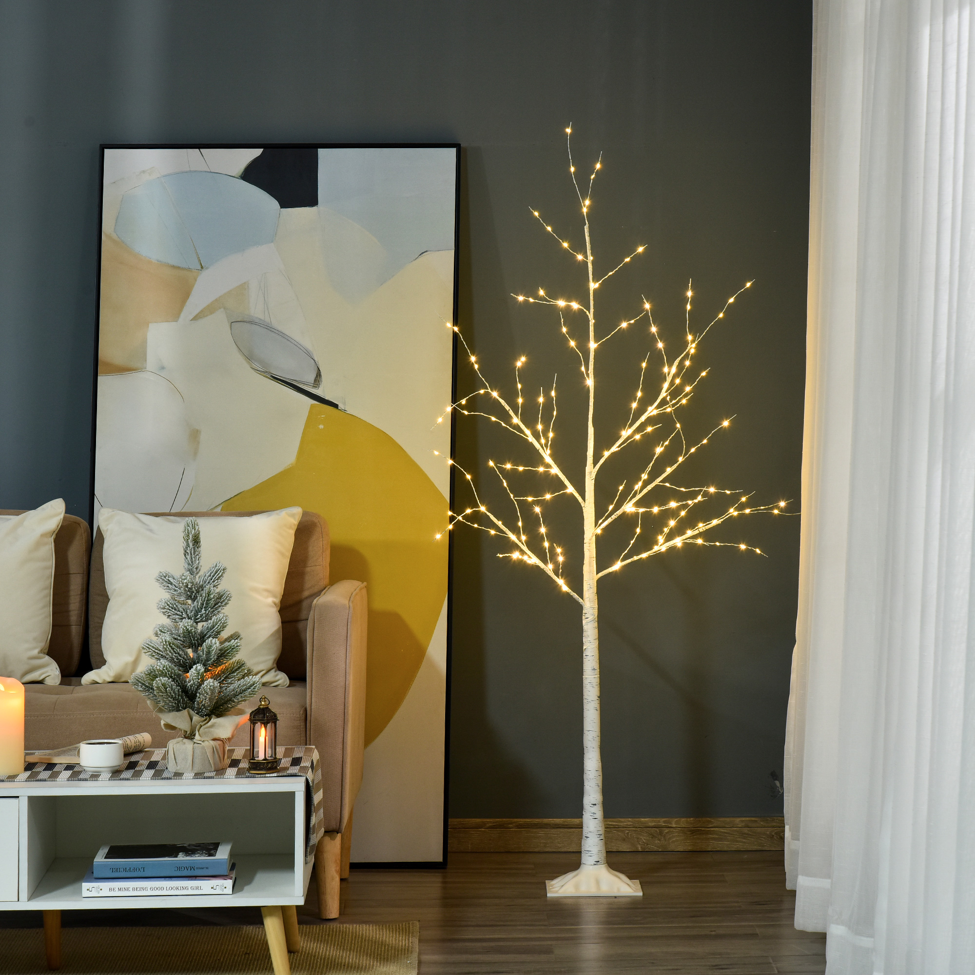 Un árbol de ramas brillante junto a un sofá beige, arte abstracto y un árbol pequeño de Navidad en una mesa.