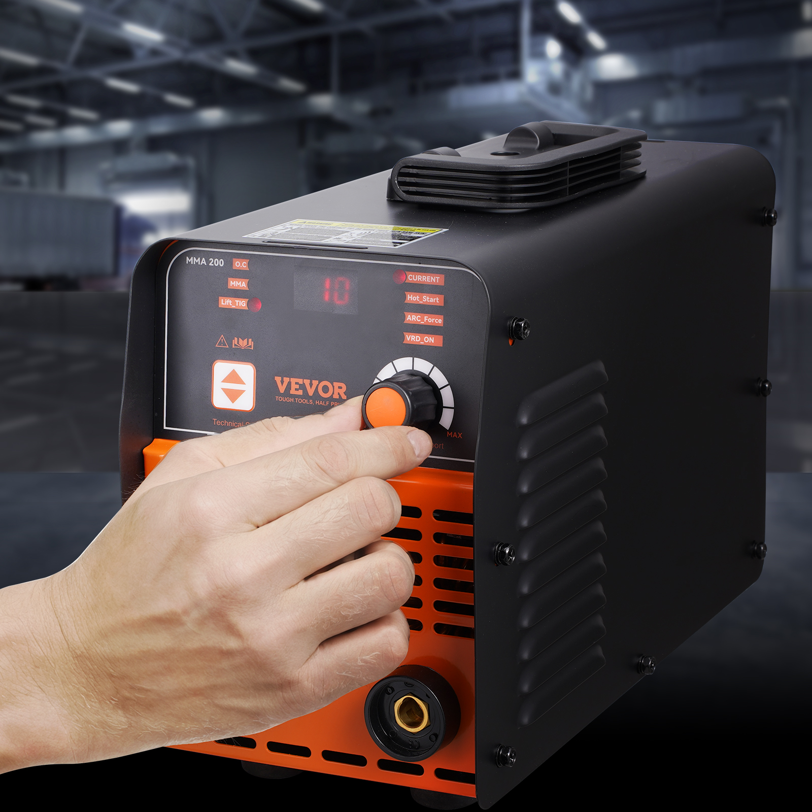 Una mano gira el dial de una máquina de soldar VEVOR negra y naranja en un taller.