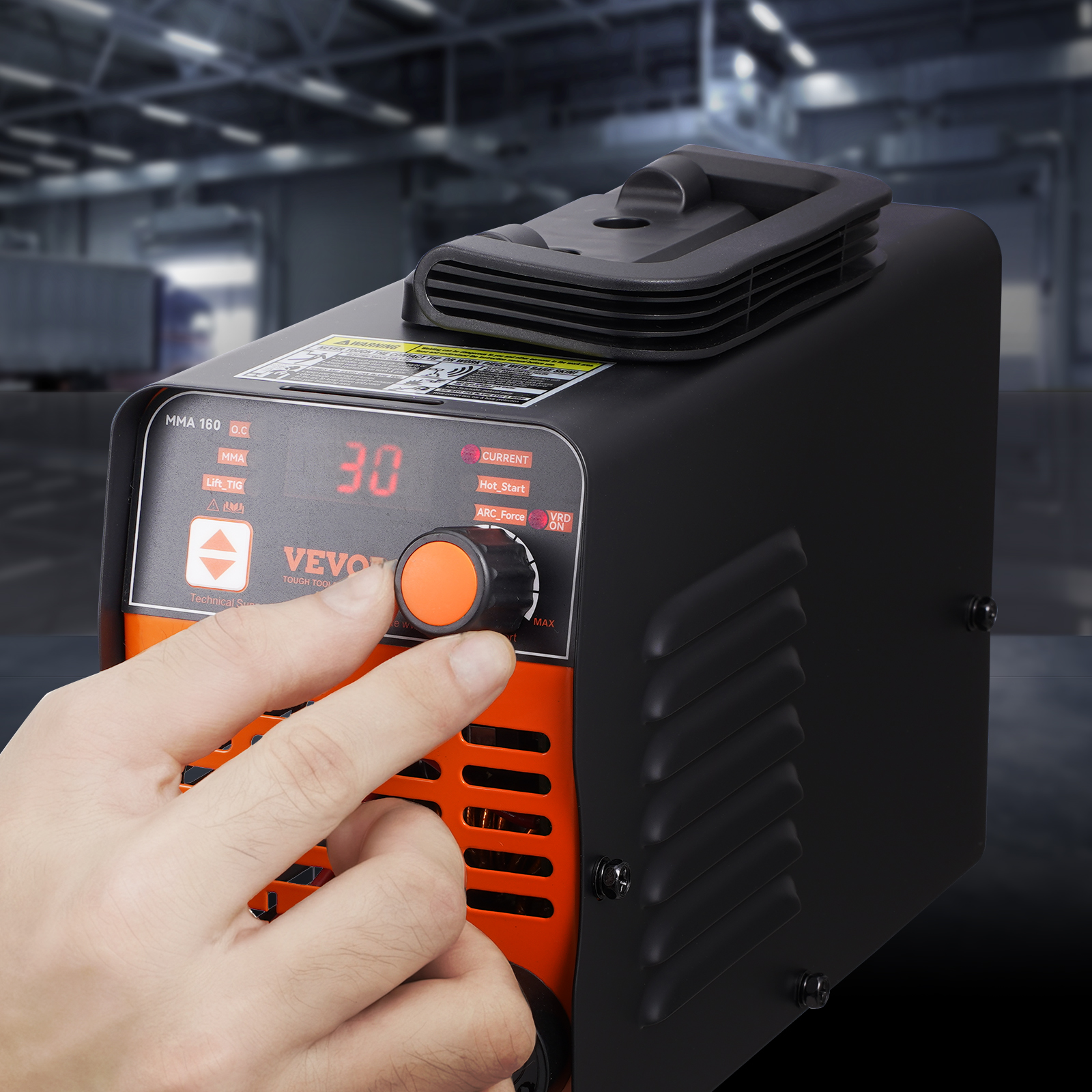 Una mano ajusta la perilla naranja en una máquina de soldar negra con pantalla digital frente a un taller.