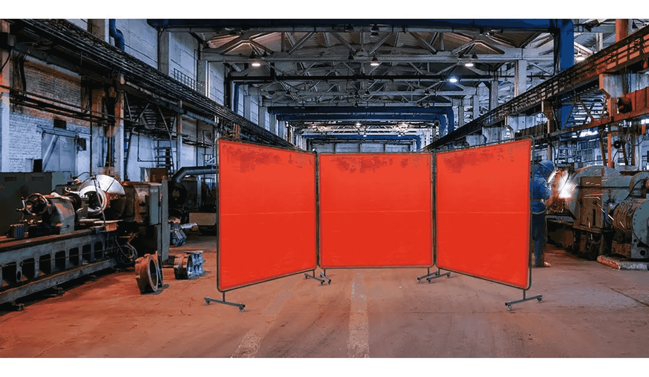 Taller industrial con mamparas de soldadura rojas y maquinaria al fondo.
