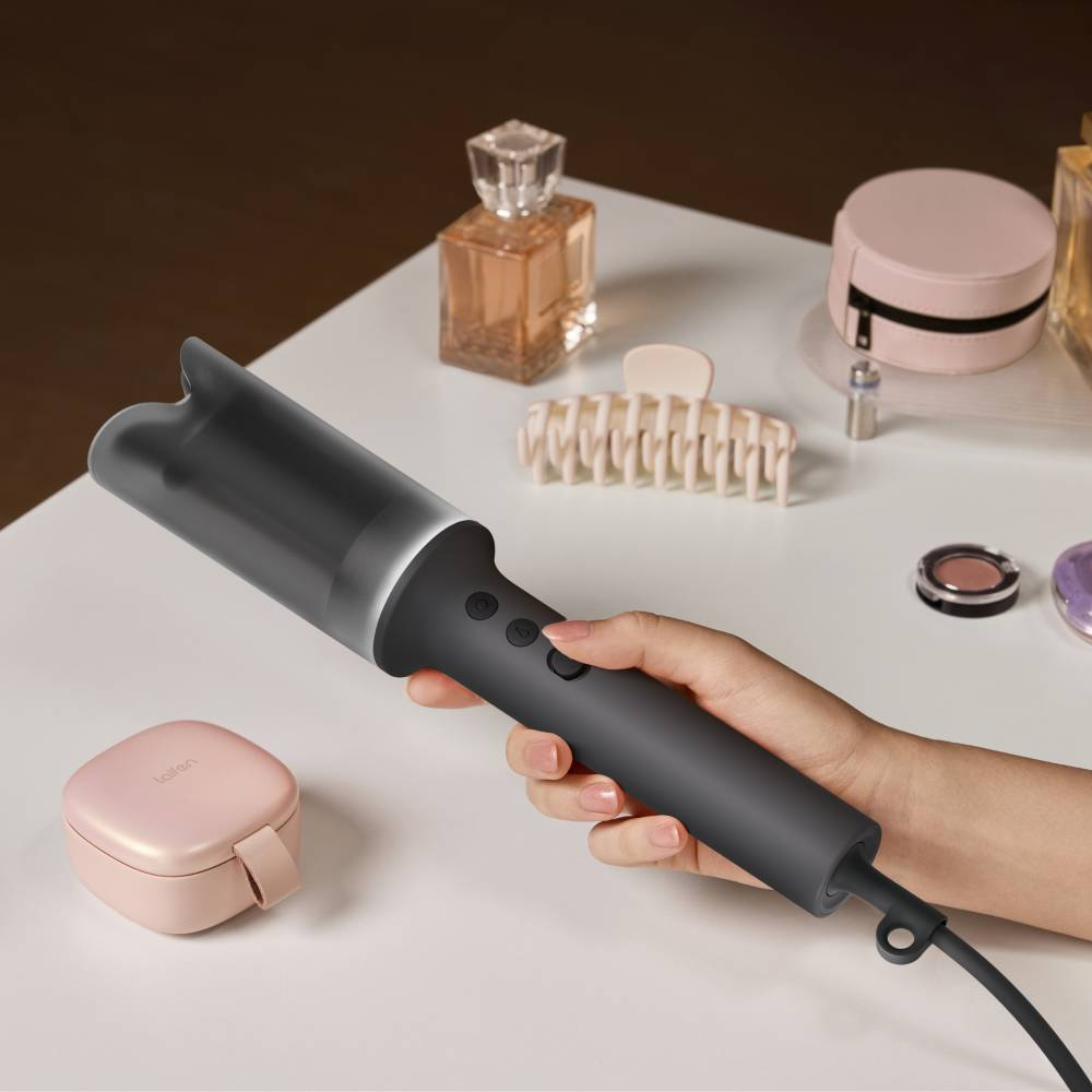 Hand hält grauen Lockenstab auf weißem Tisch mit Kosmetik und Parfum.