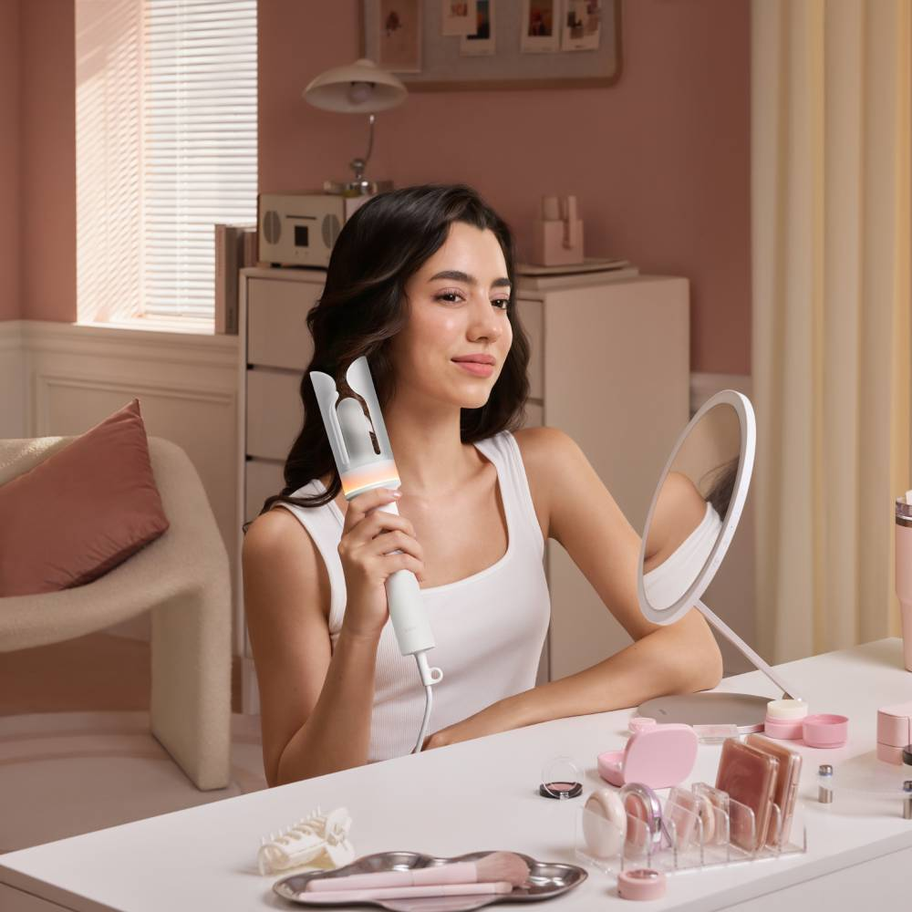 Une femme à sa coiffeuse utilise un fer à friser blanc devant un miroir.