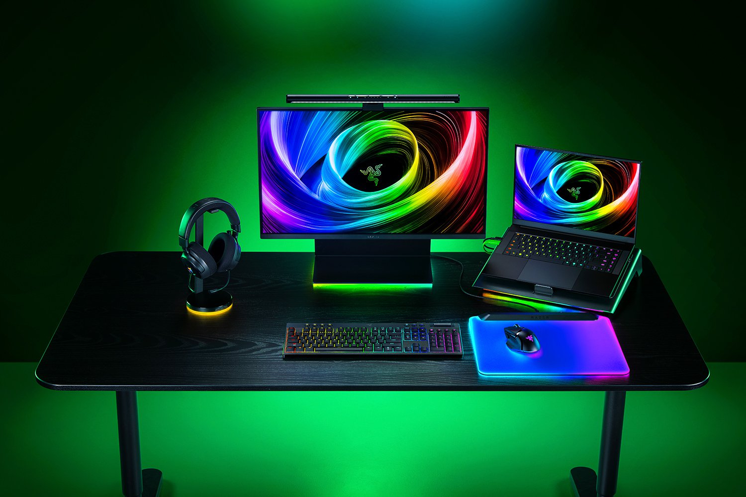Postazione gaming RGB su scrivania nera con monitor, laptop, tastiera, mouse, tappetino, cuffie e illuminazione.